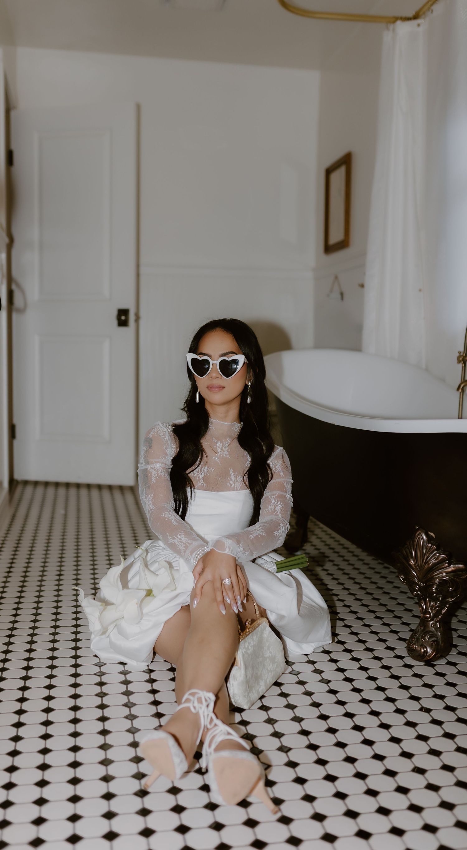Bride in white lace dress and heart sunglasses poses on checkered bathroom floor near clawfoot tub.