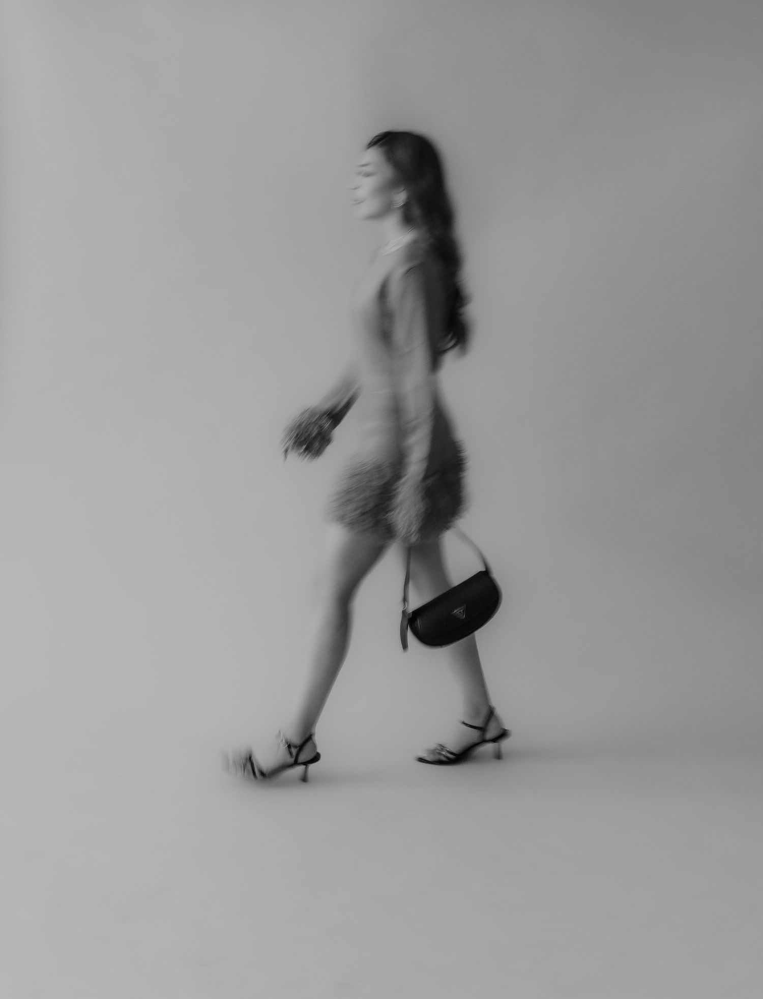 Blurred black and white photo of a woman walking in a feathered dress and heels carrying a small handbag.