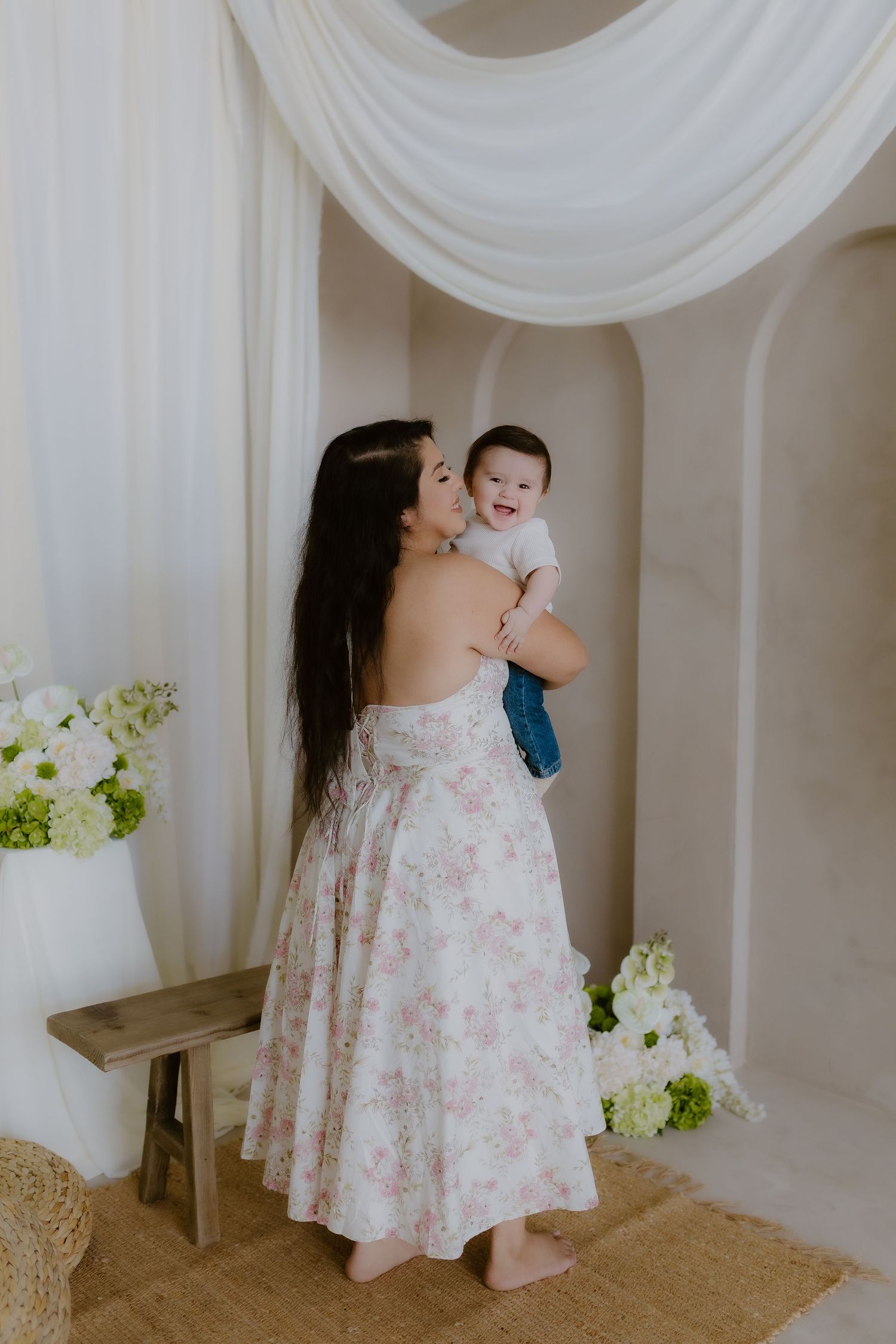 Mother in floral dress lovingly holds and kisses her smiling baby in a bright white studio setting.