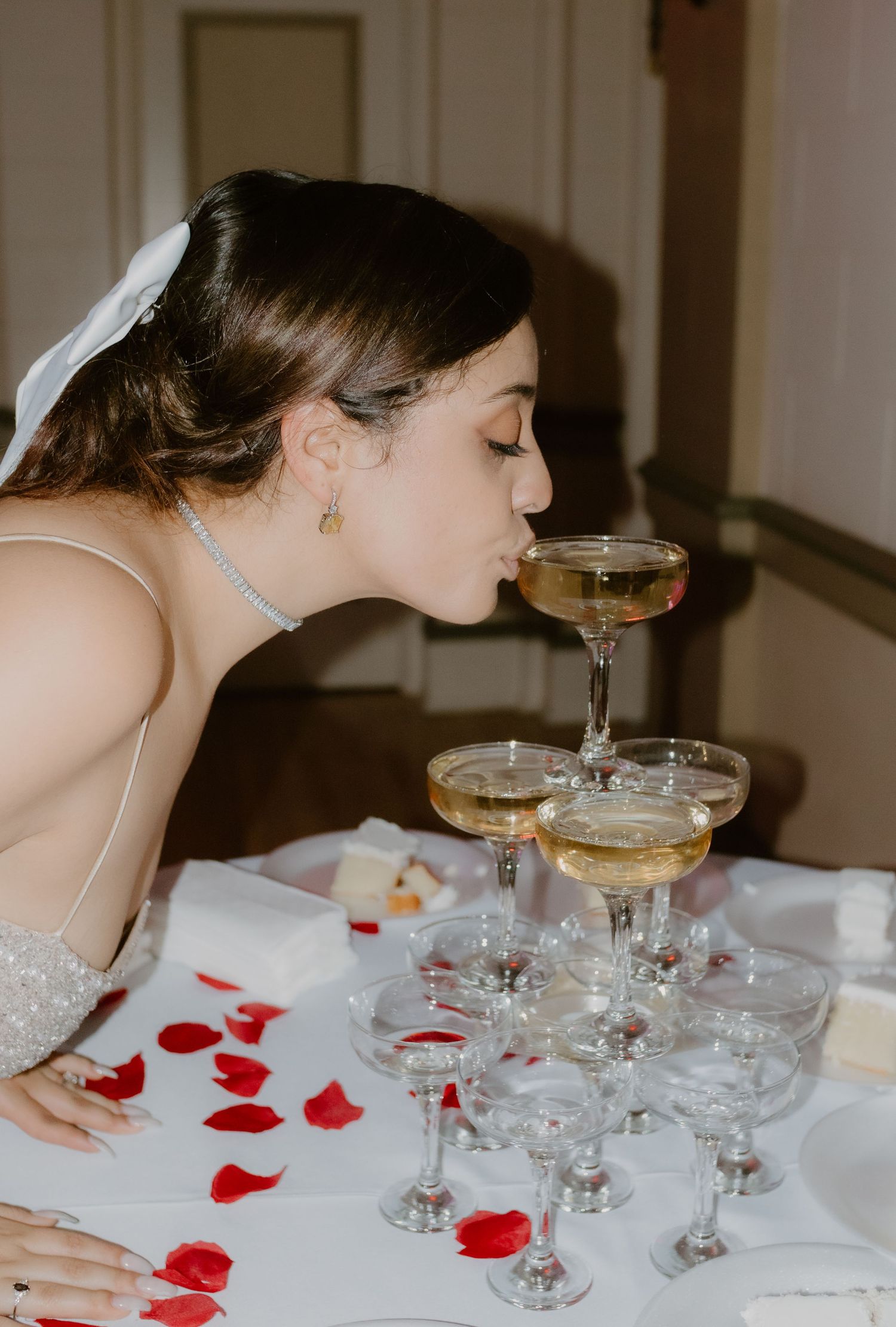 Bride in white wedding gown sipping champagne directly from a glass in a champagne tower at her wedding reception.