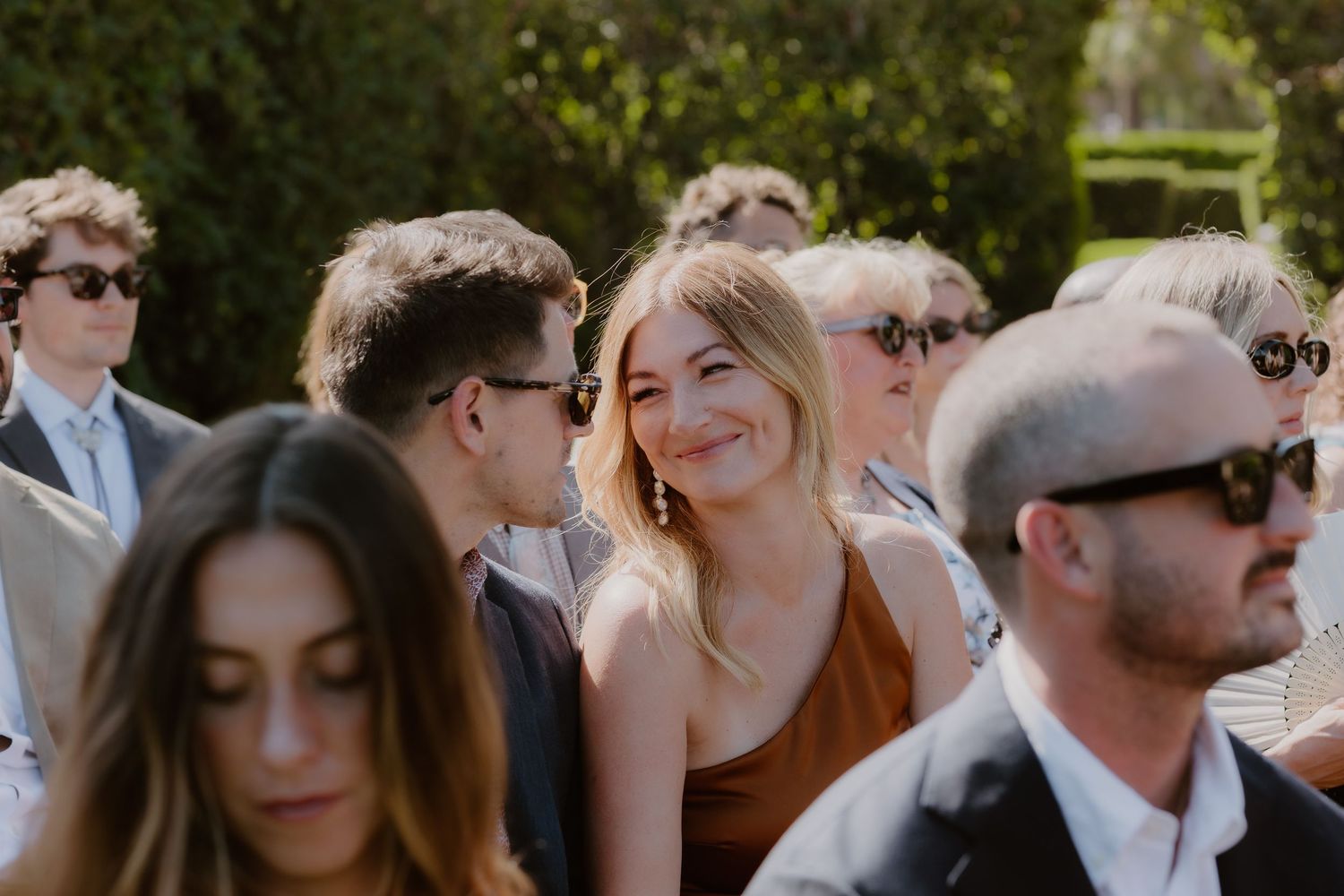 Smiling female wedding guest in a brown dress stands out among other guests at an outdoor wedding reception.