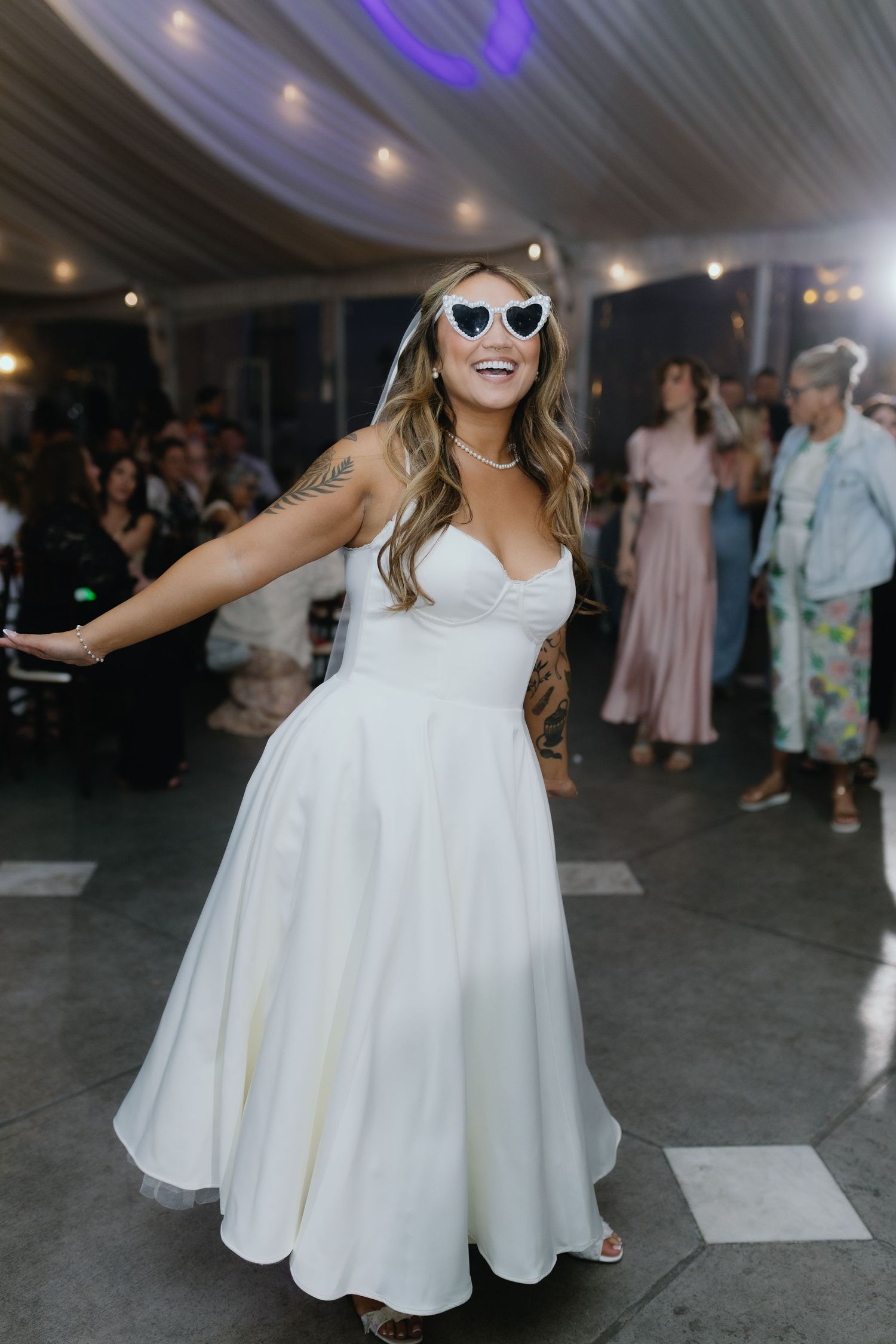 Joyful bride dancing in short white dress wearing heart-shaped sunglasses at wedding reception.