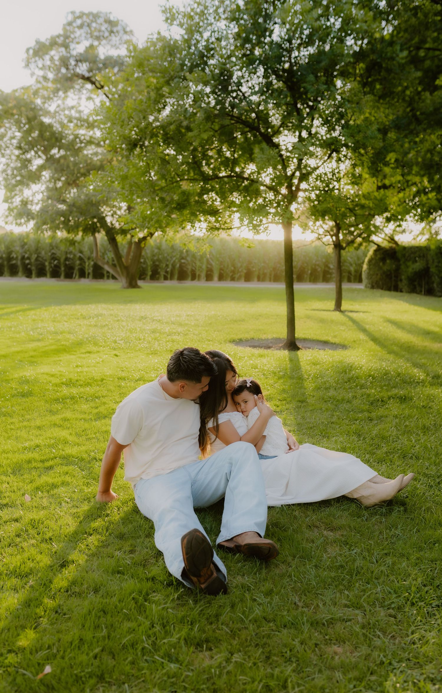 Family of three sitting together on green grass in golden sunlight, parents gazing lovingly at young daughter.