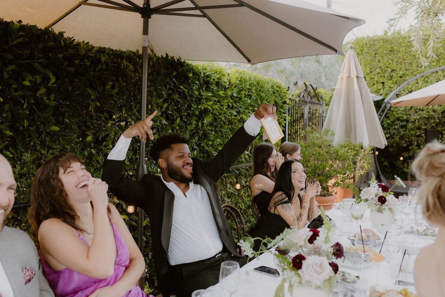 Wedding guest raises a toast and laughs joyfully at an outdoor garden reception table with floral centerpieces.