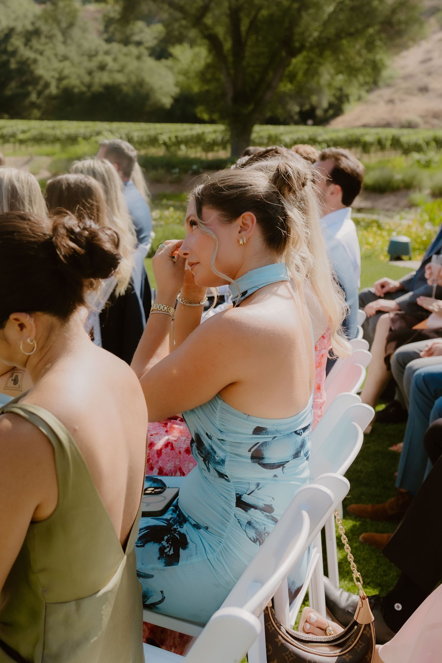 Emotional wedding guest in floral blue dress wipes tears during outdoor garden ceremony.