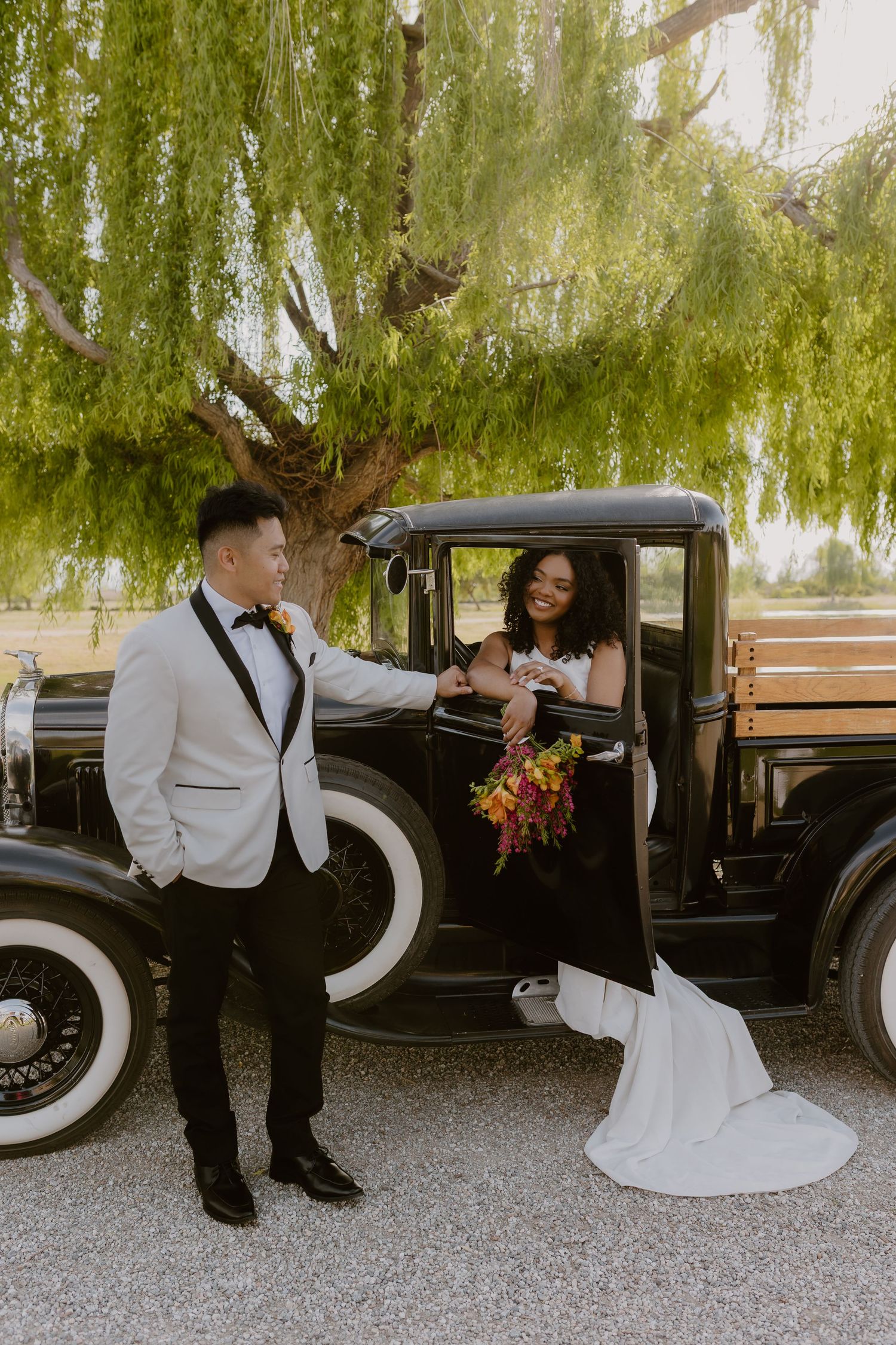 Groom in white tuxedo helps bride in sleek white gown exit a vintage black antique truck under a willow tree.