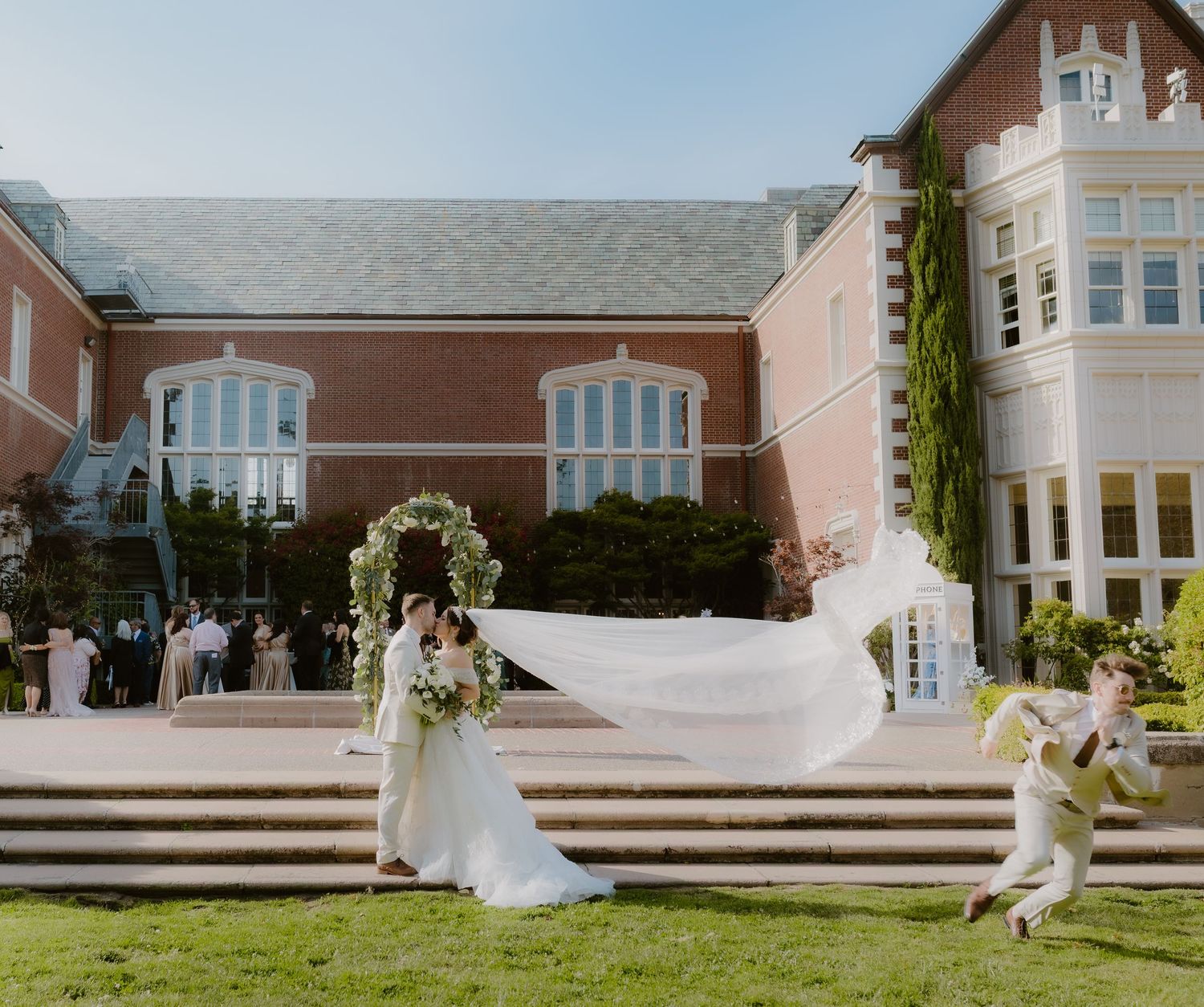 Bride and groom share romantic kiss outdoors as wedding veil flows in wind near brick venue.