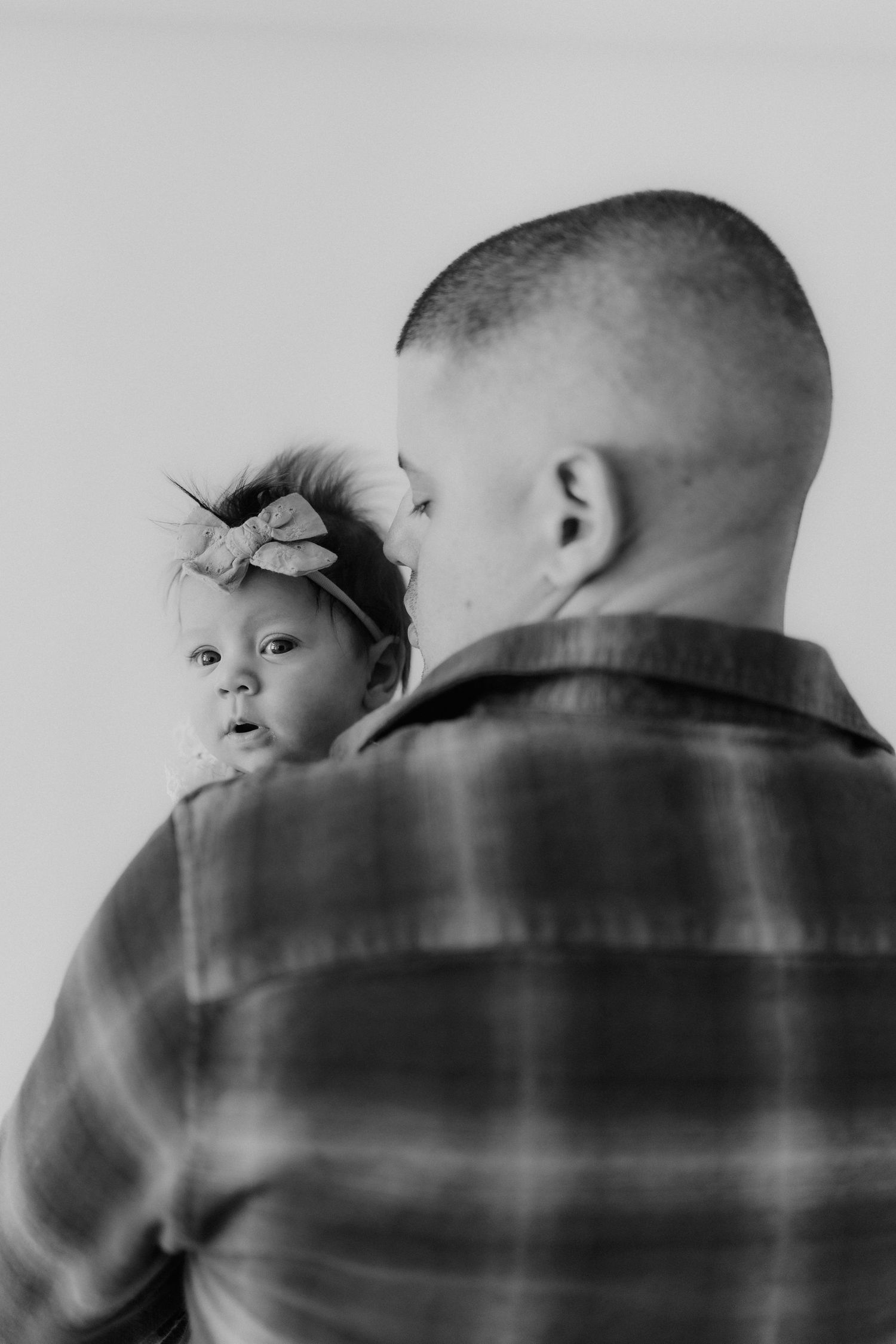 Black and white photo of a father in plaid shirt holding his newborn baby girl with bow headband.