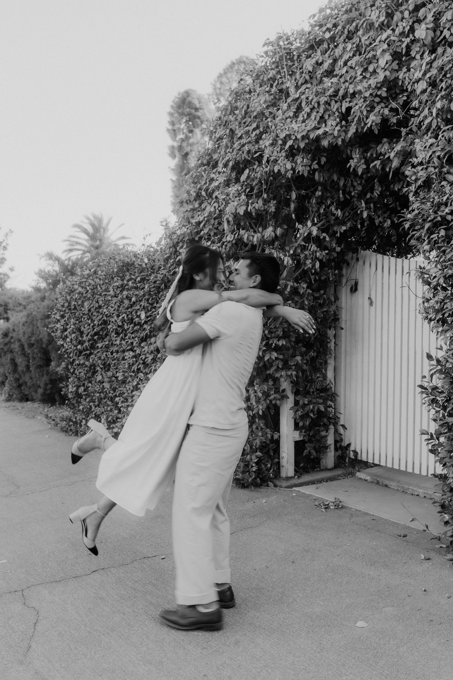 Black and white photo of couple embracing and lifting near ivy-covered white gate.