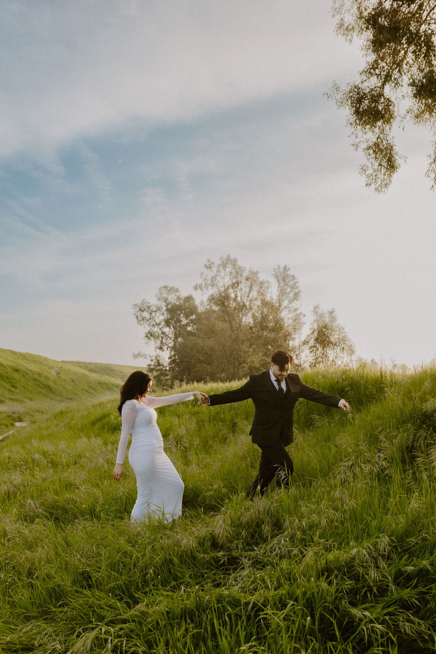 Bride and groom playfully walking up grassy hillside holding hands under blue sky.