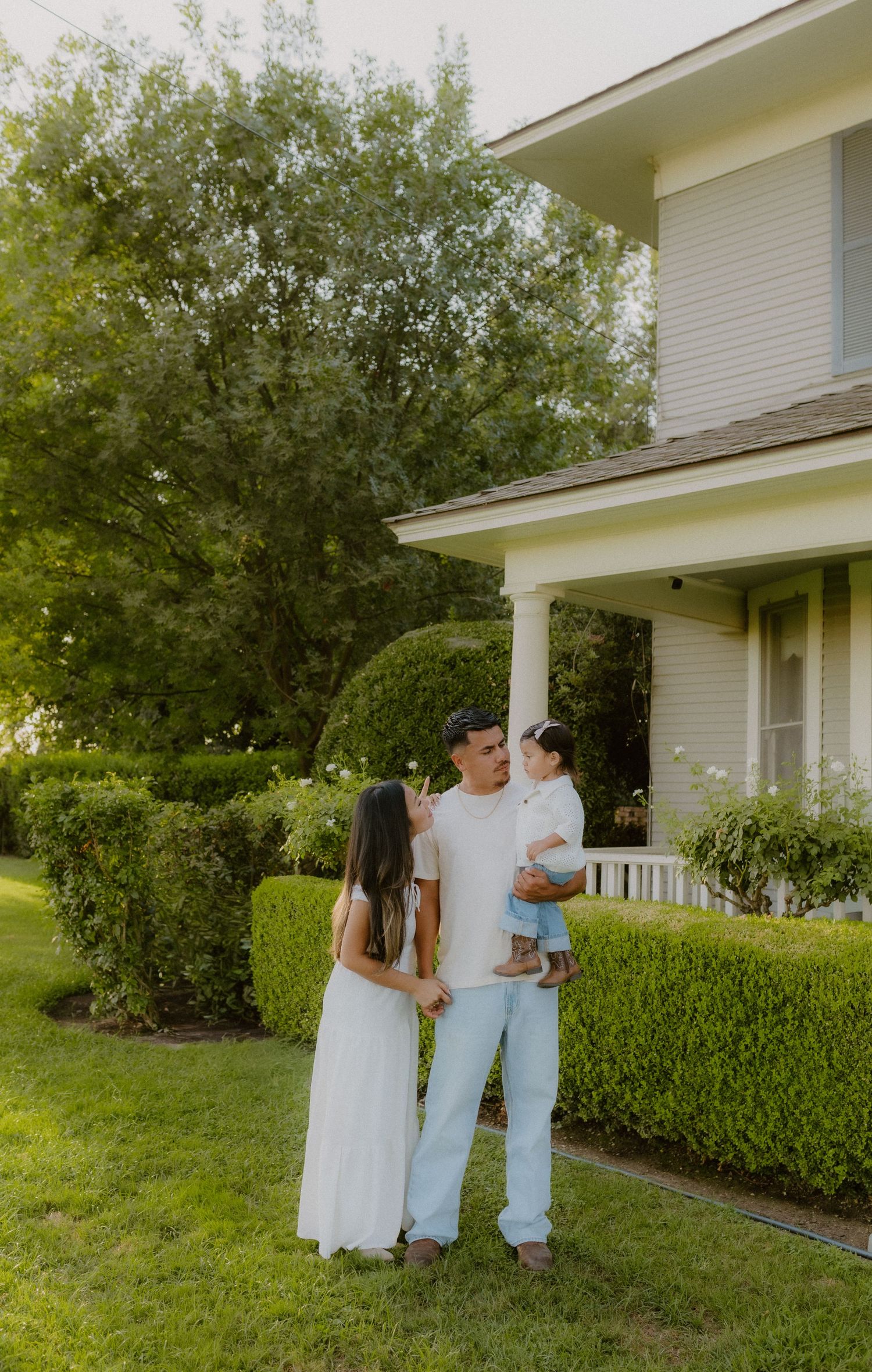 Young family of three posing in front of a charming white farmhouse surrounded by green hedges.