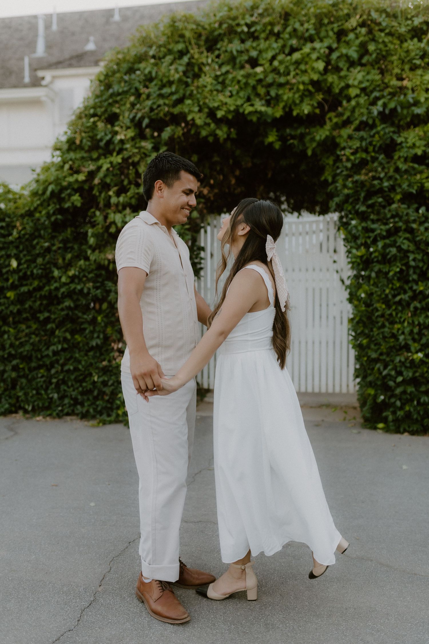 A couple in white and beige outfits hold hands and smile at each other in front of a lush green hedge.