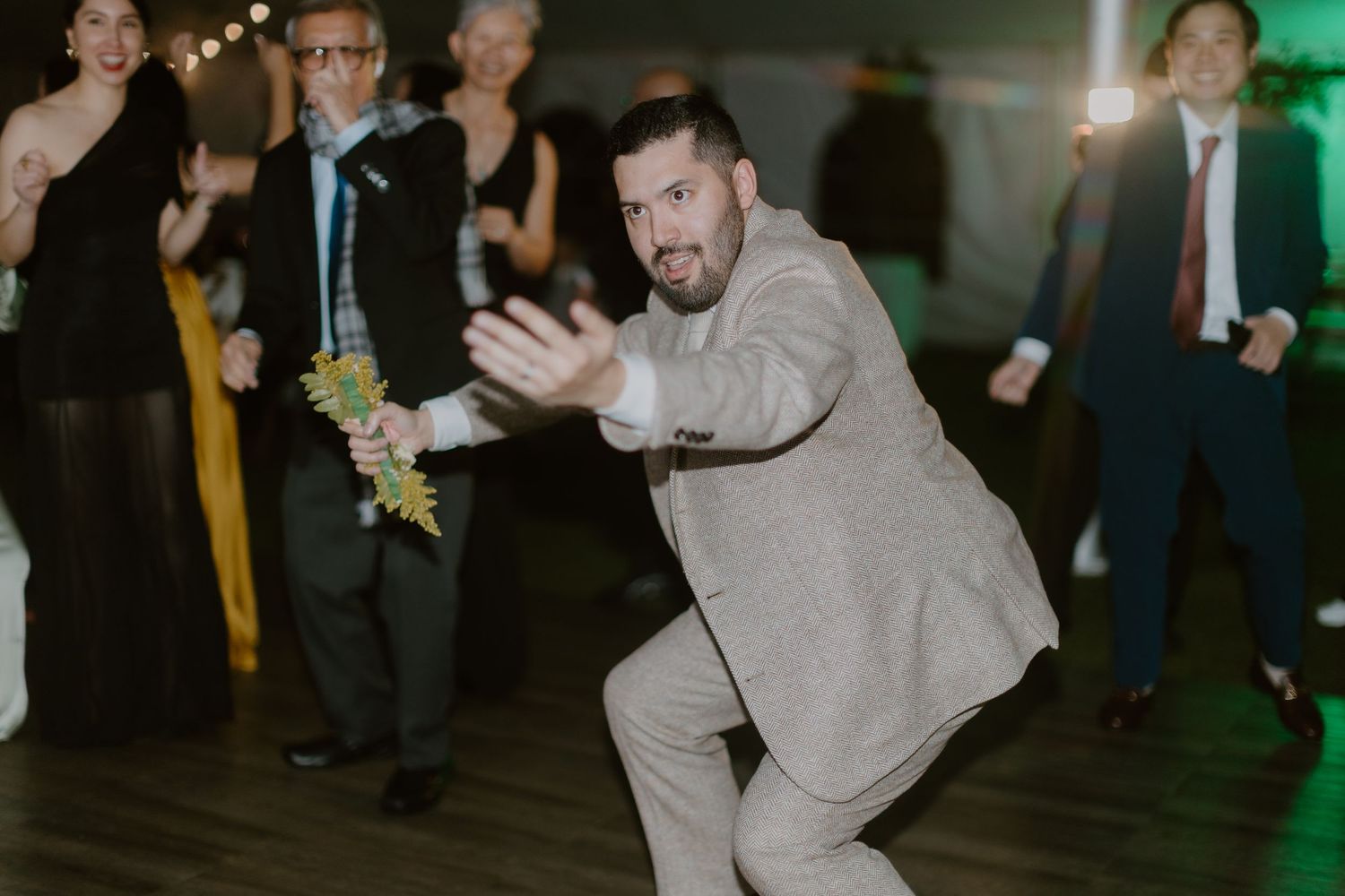 Groom in tan suit energetically dancing at wedding reception with guests in background.