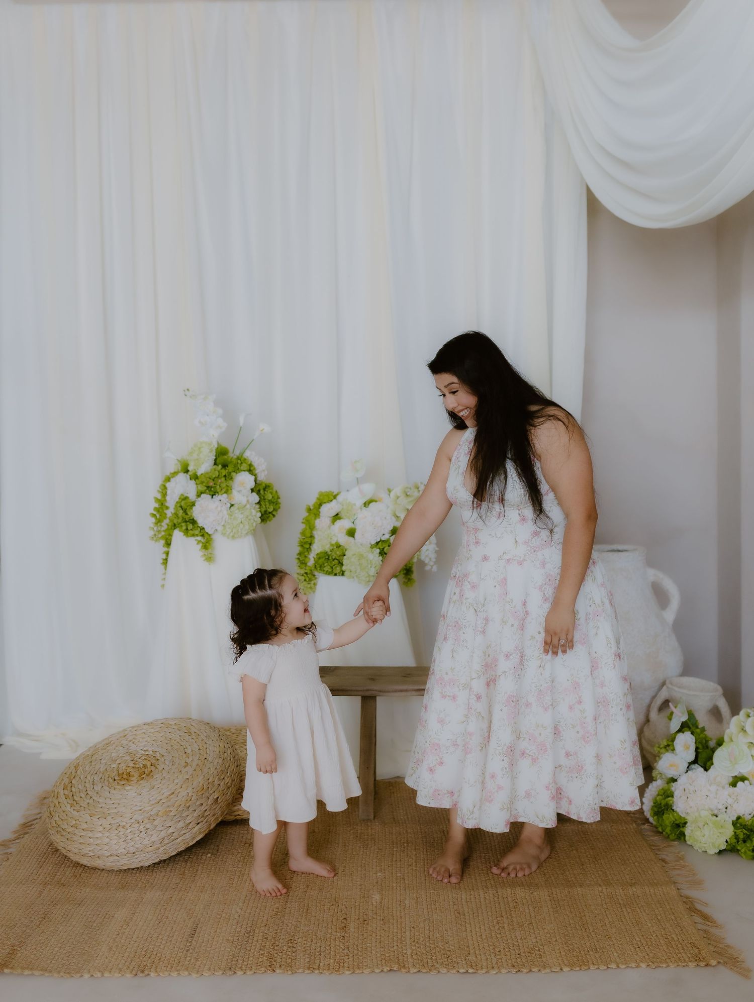Mother in floral dress holding hands with toddler daughter in white dress in bright studio setting. mini session Tulare california
