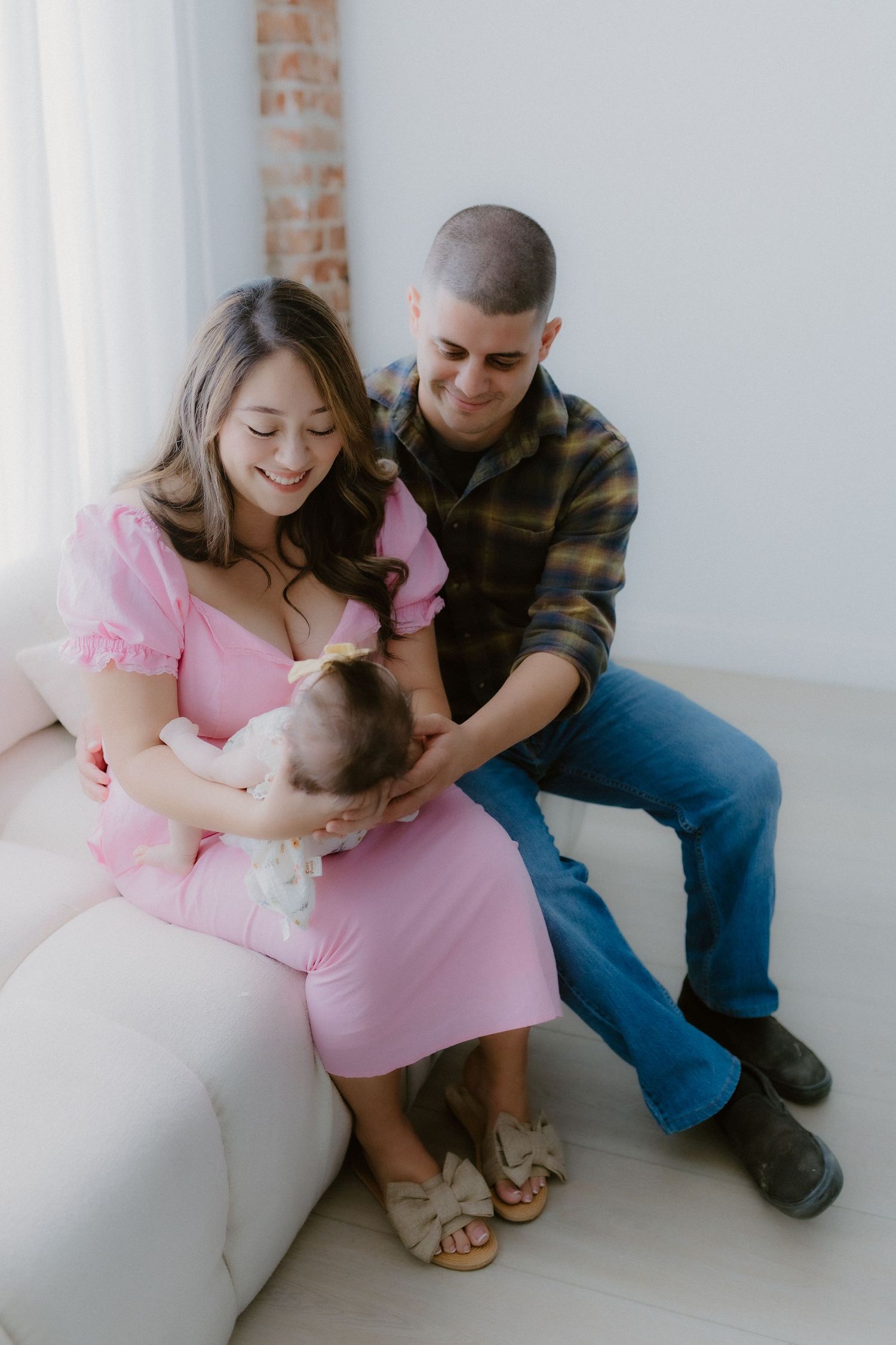 Happy young family in pink and plaid sitting on white couch with newborn baby girl.