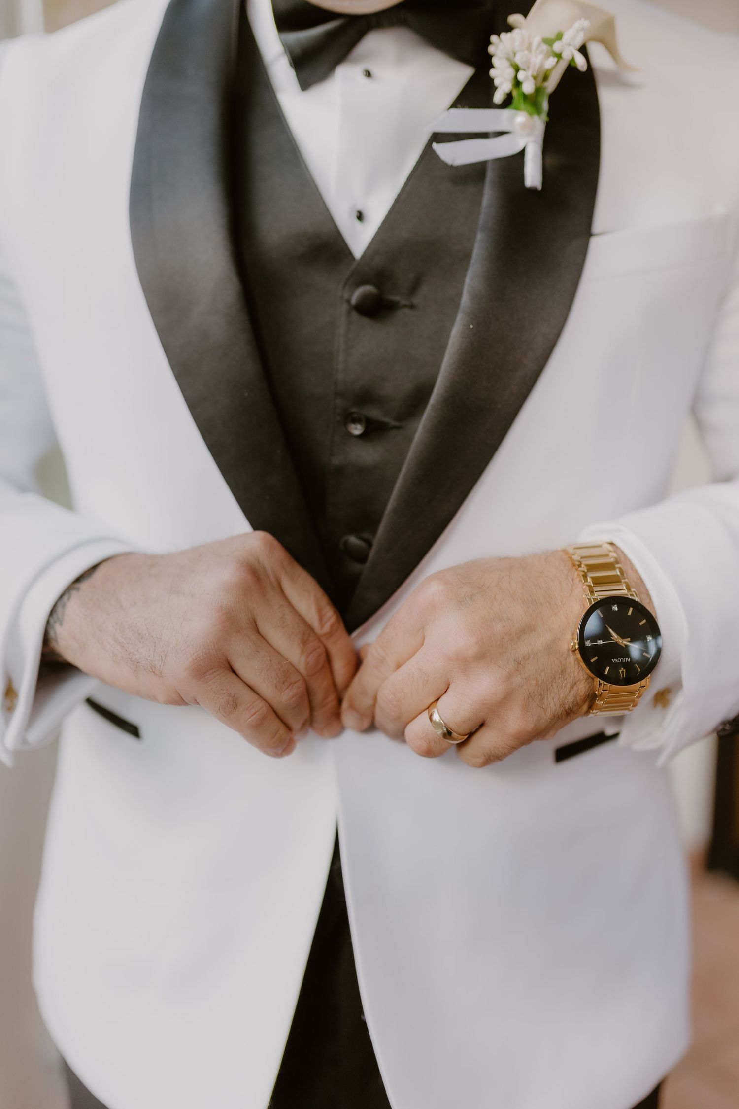 Groom in white tuxedo jacket with black lapels adjusts buttons, wearing gold watch and floral boutonniere.