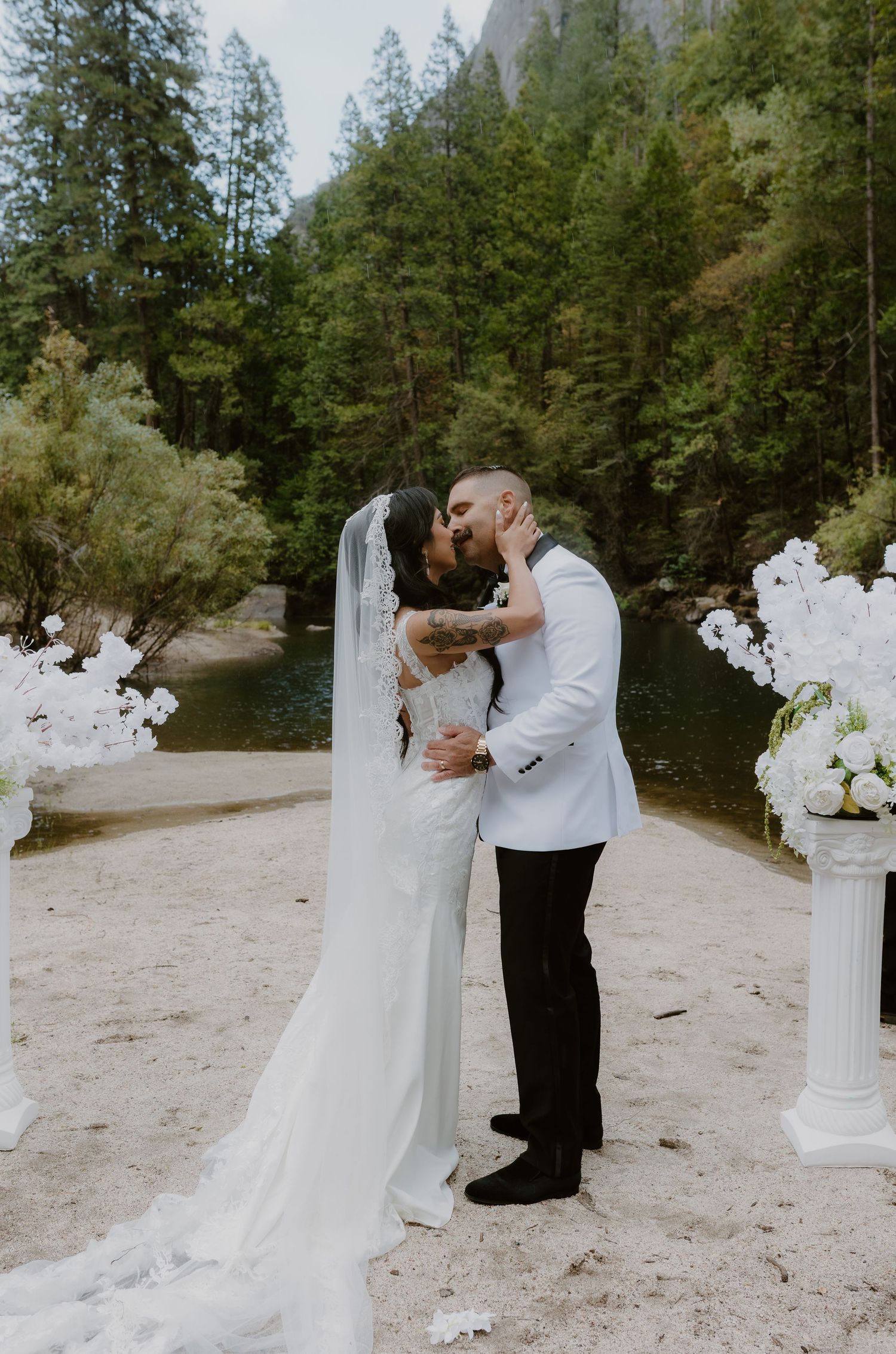 Bride and groom sharing first kiss at outdoor Yosemite wedding ceremony surrounded by white floral arrangements.