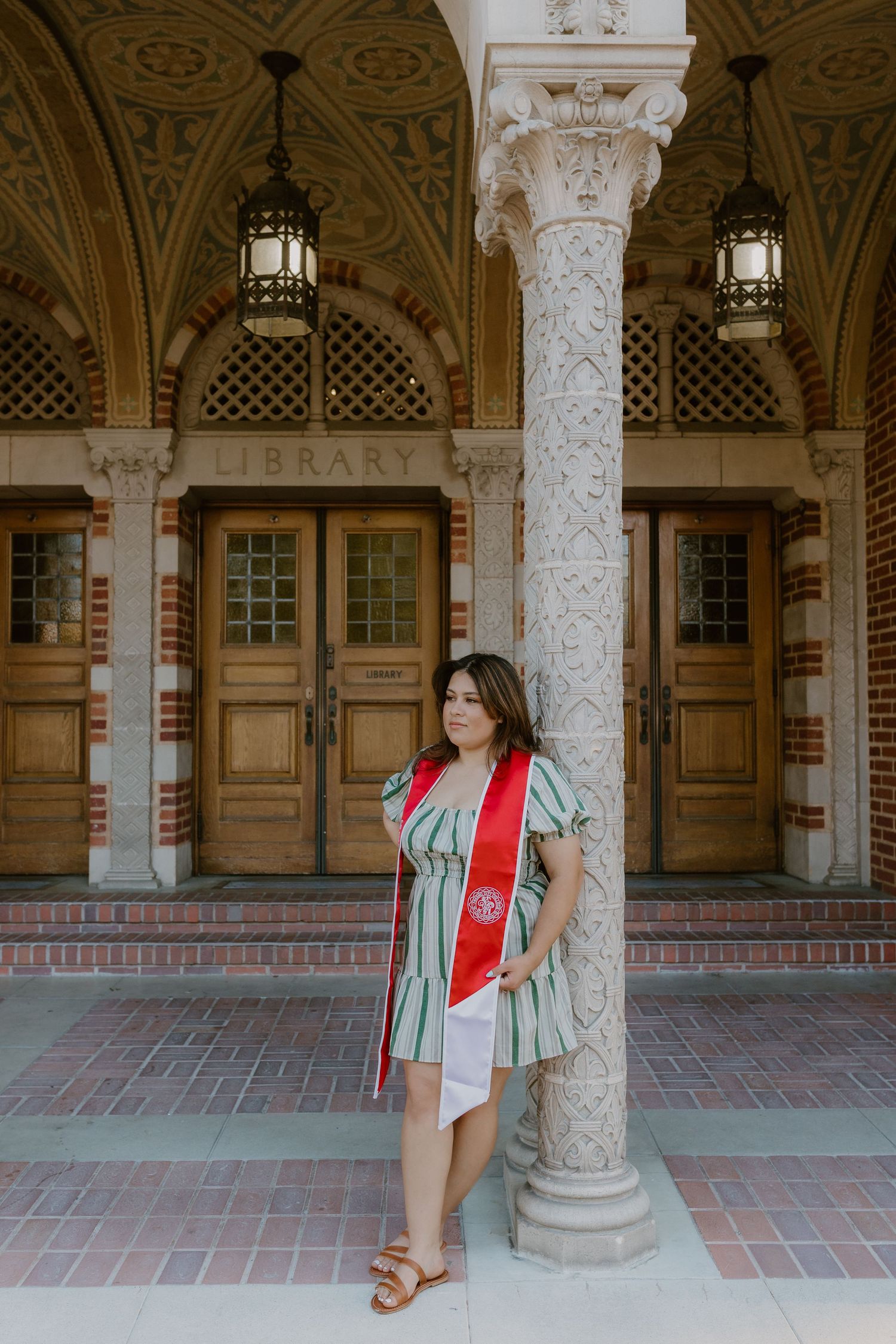 Graduate in striped dress with red sash leans against ornate column outside university library entrance. Fresno Pacific University