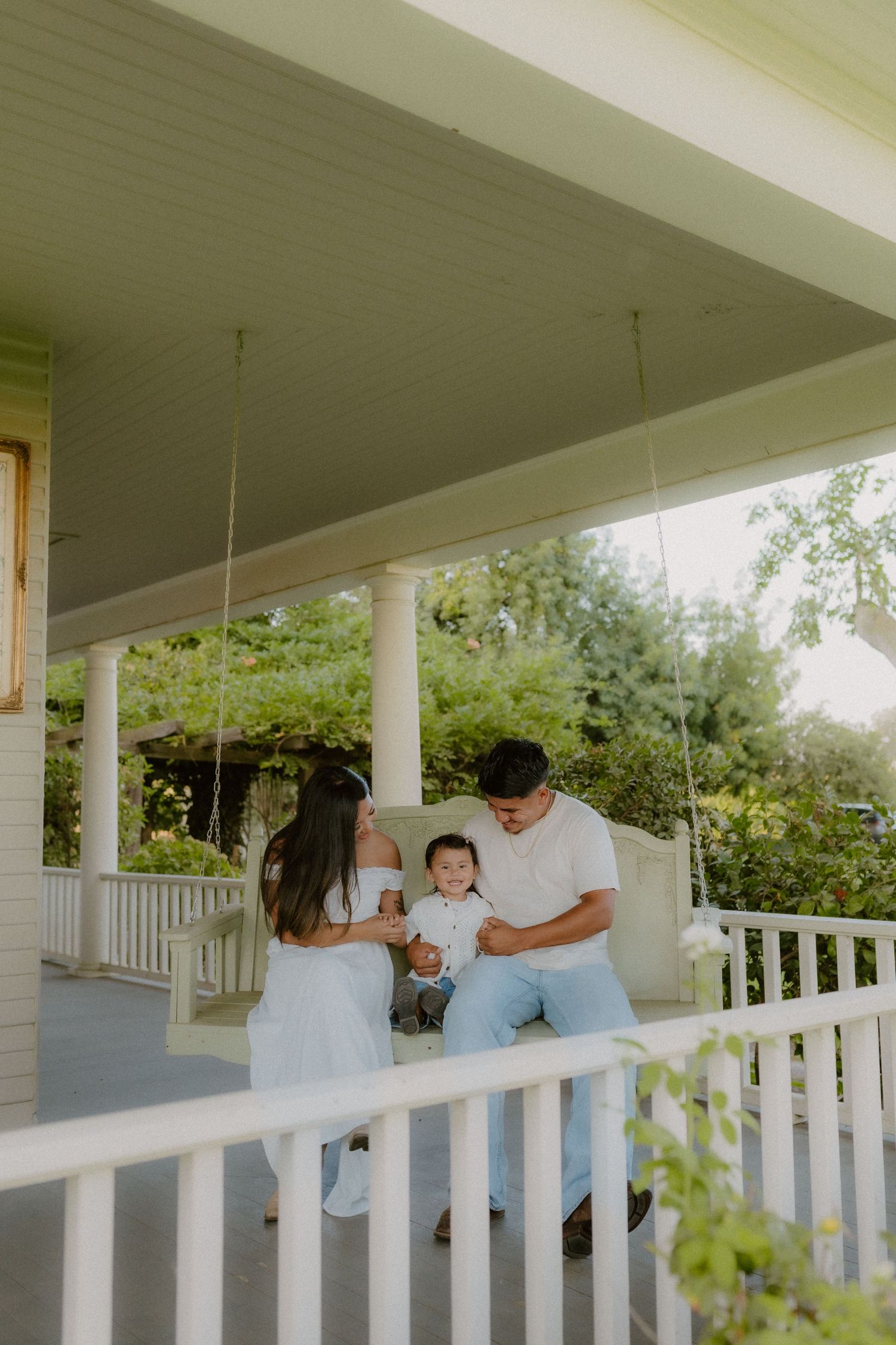 A young family sits on a white porch swing, parents smiling at their baby on a charming home's front porch.