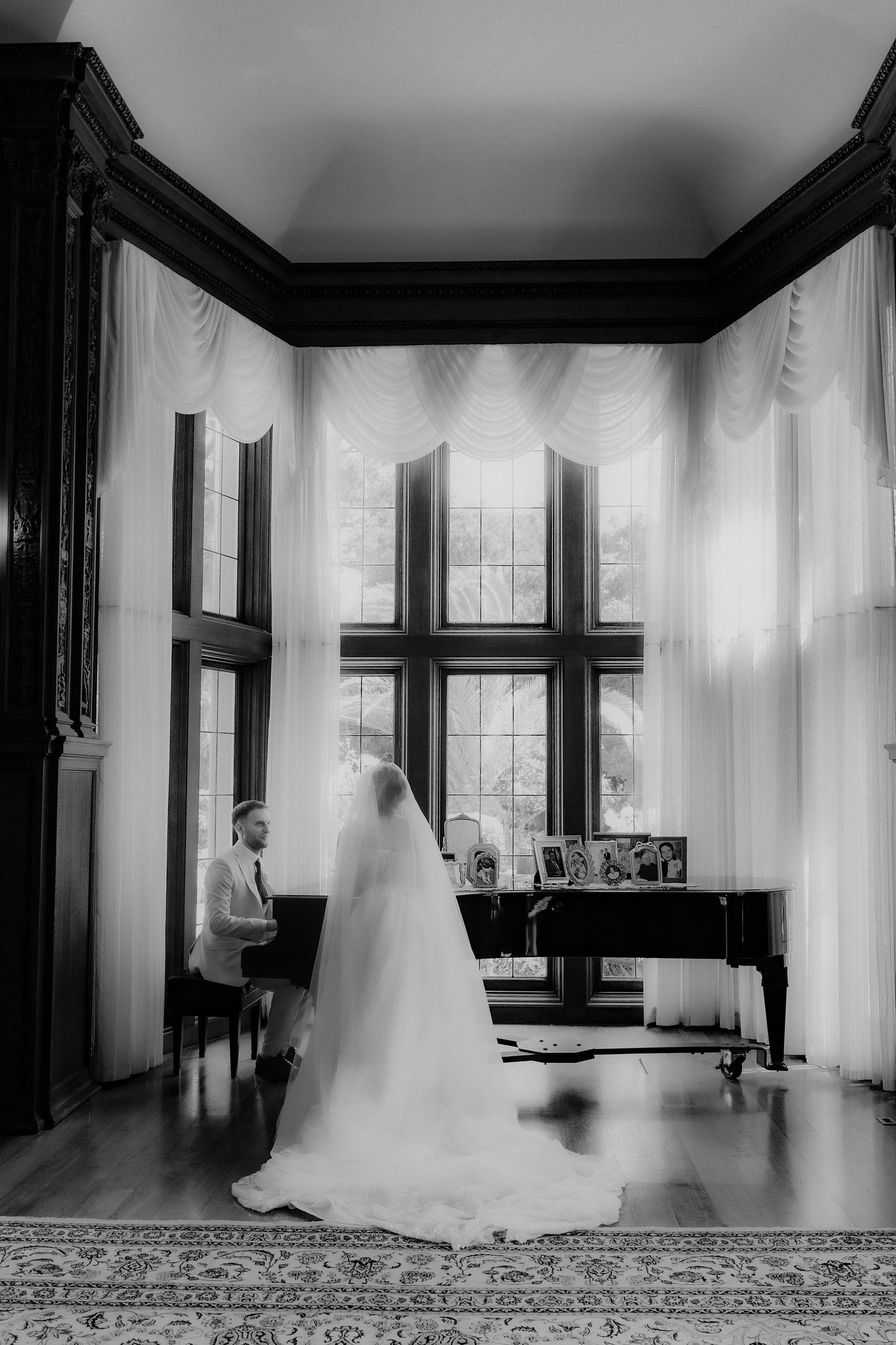 Black and white photo of bride in cathedral veil standing by grand piano near tall windows.