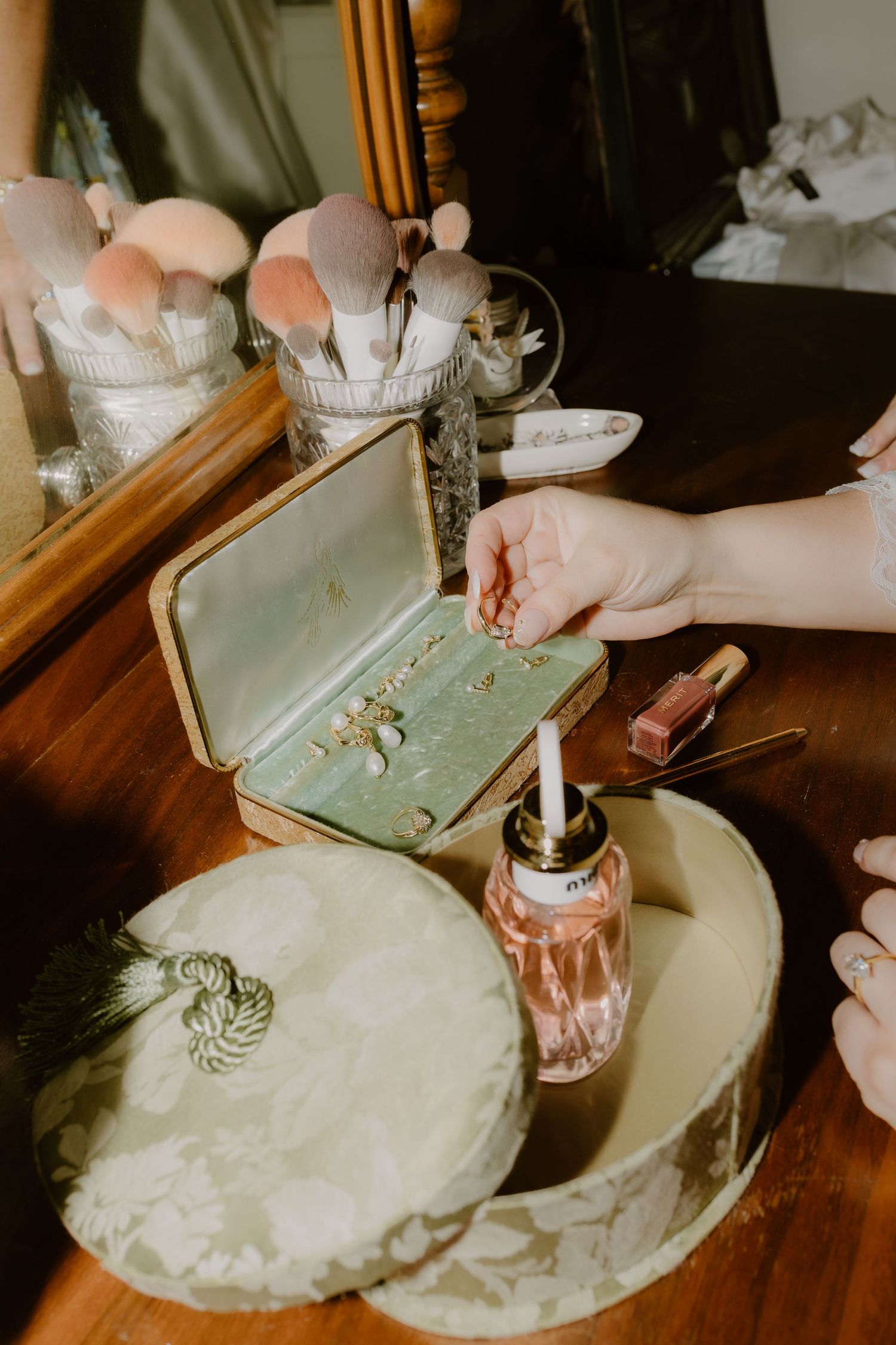 Bride selecting jewelry from vintage green velvet box at wooden vanity with makeup brushes.