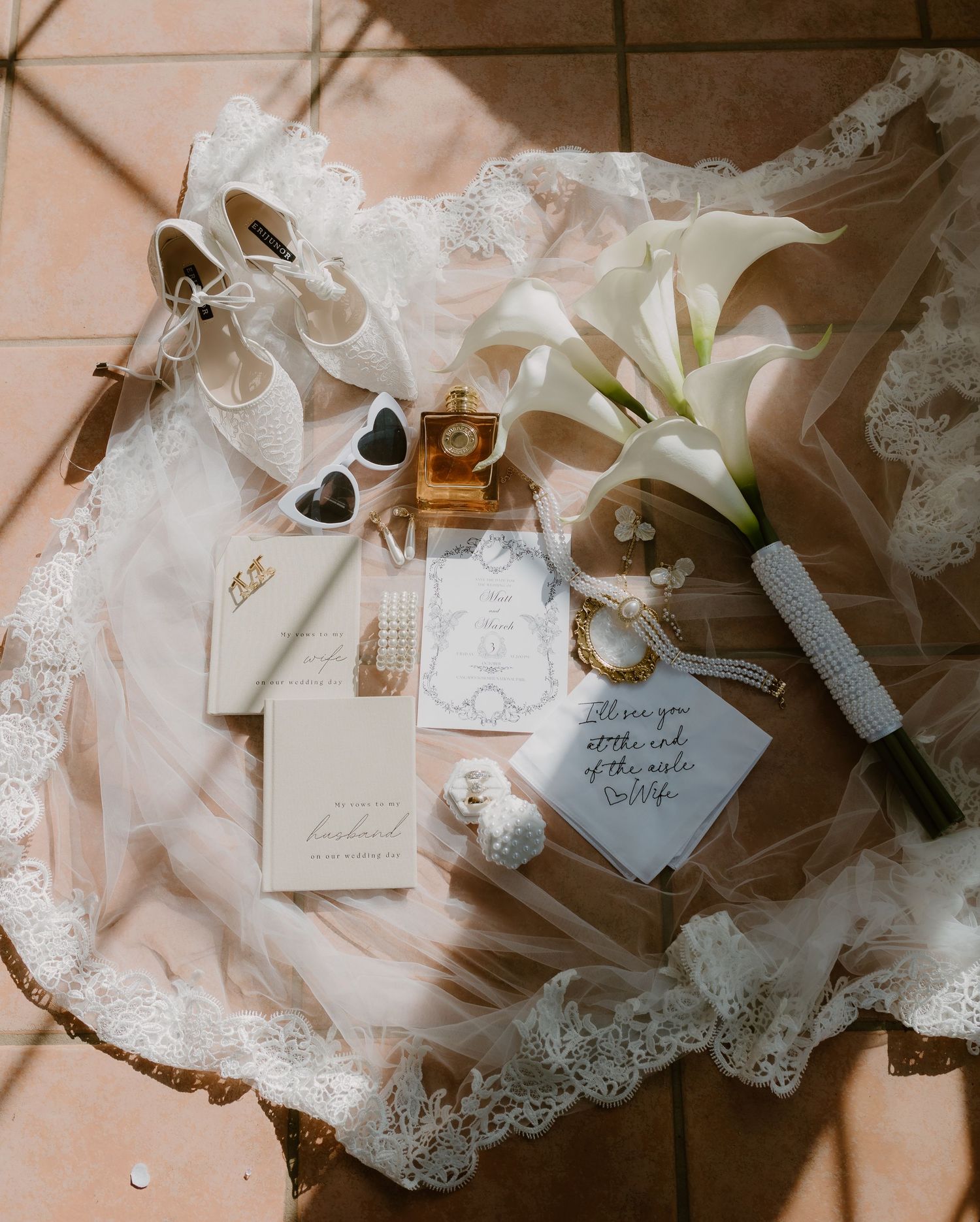 Flat lay of bridal accessories including white heels, calla lily bouquet, invitations, and jewelry on lace veil.