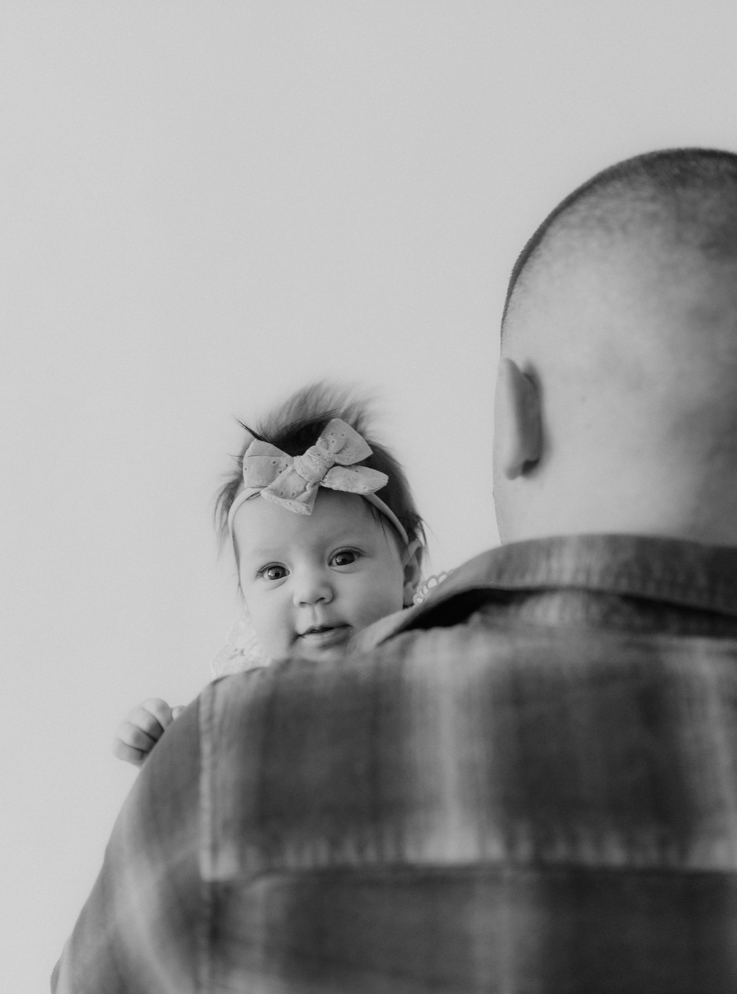 Black and white image of a baby girl in a bow headband peeking over her fathers plaid-shirted shoulder.