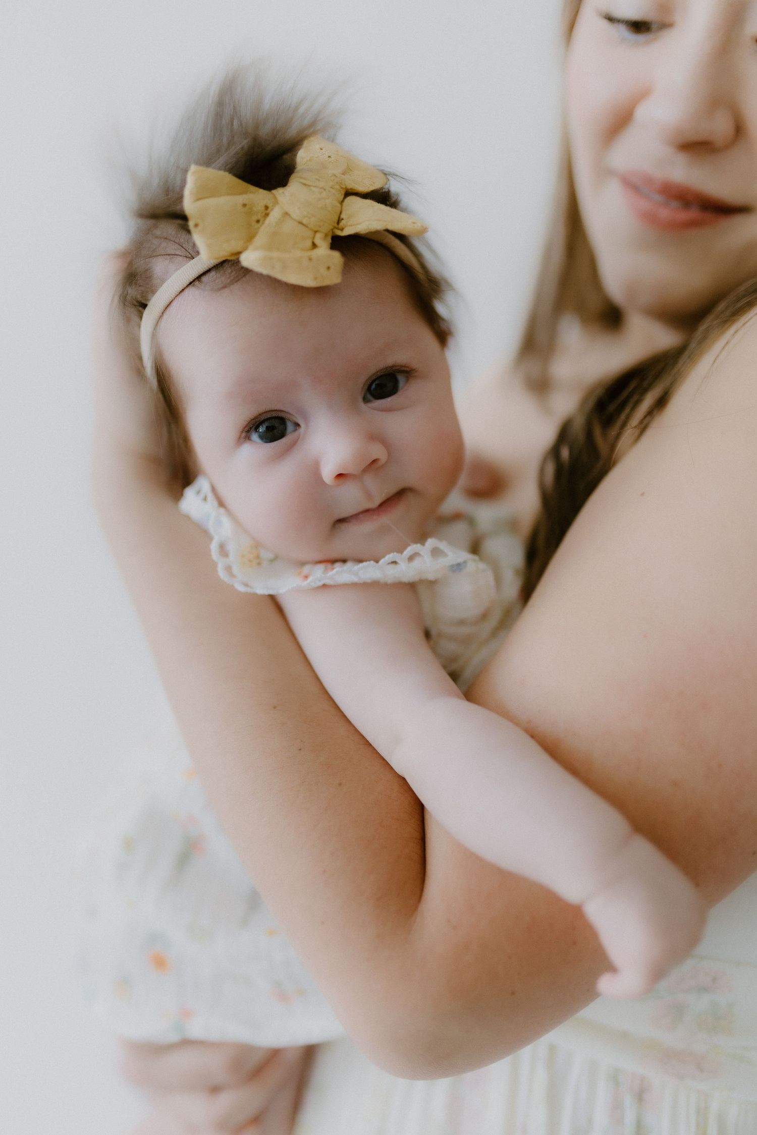 Close-up portrait of a newborn baby girl with a gold bow headband looking at camera while held by mother.