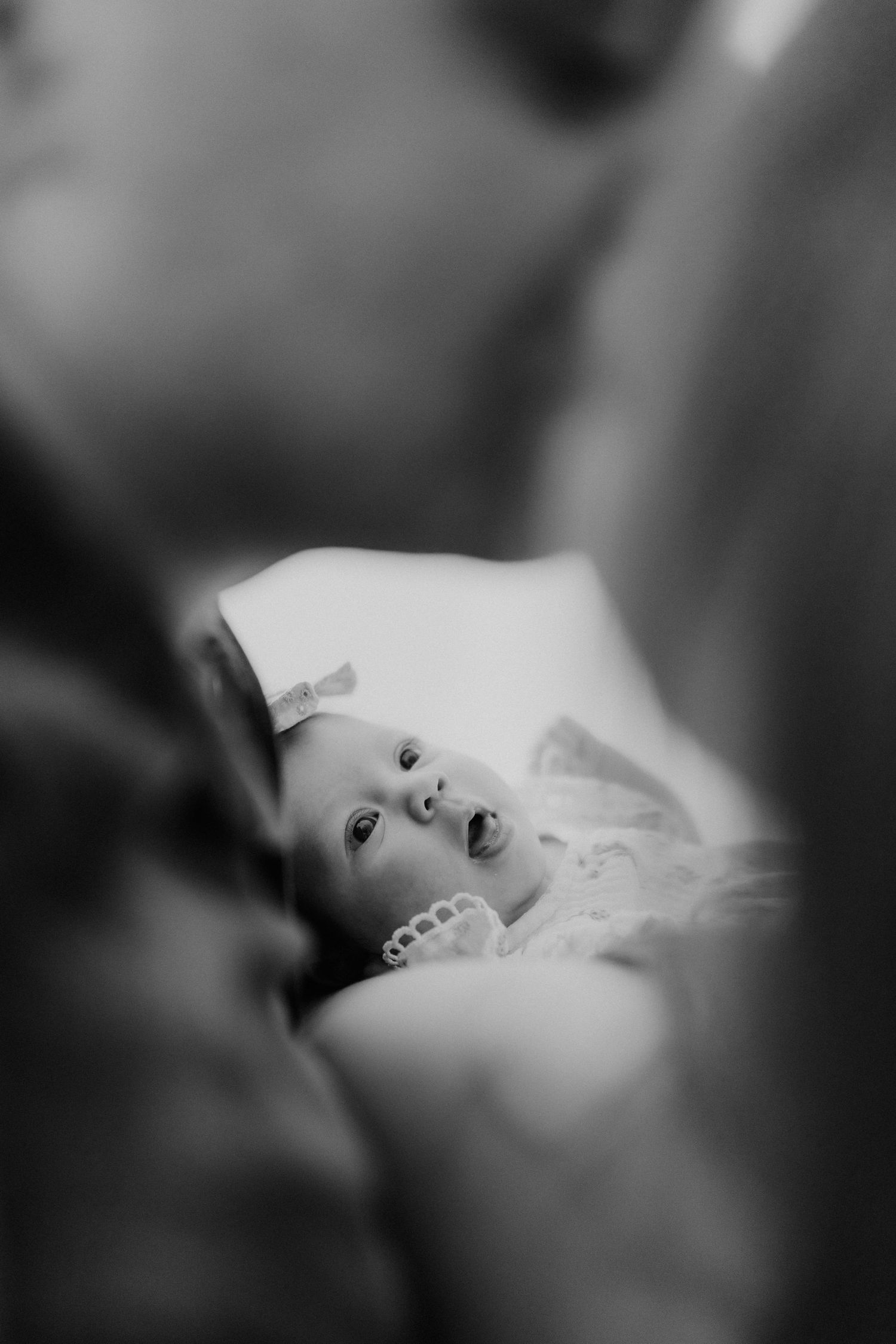 Black and white photo of a newborn baby girl with bow looking up between two blurred parents.