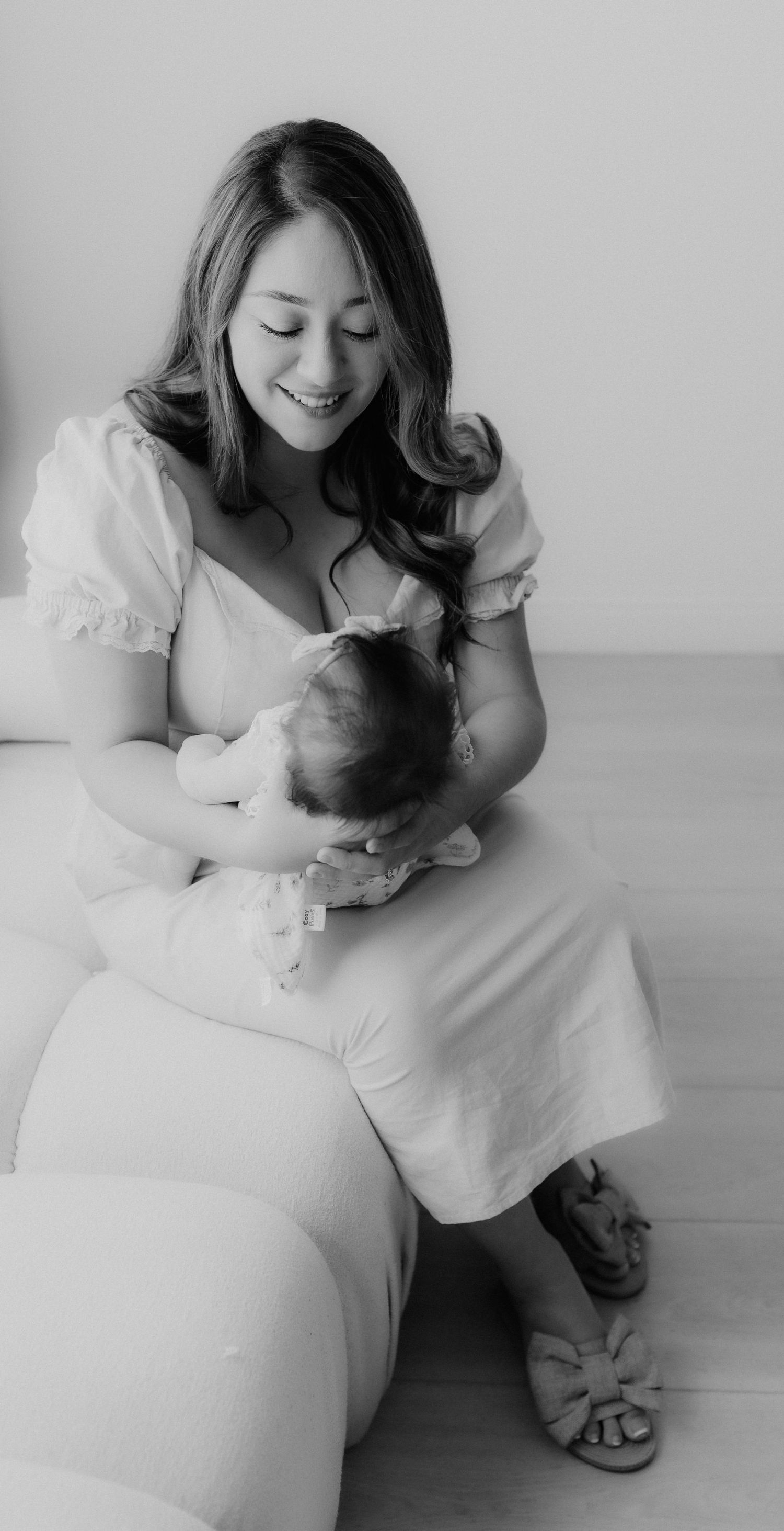 Smiling mother in white dress holding newborn baby while seated on white couch in black and white photo.