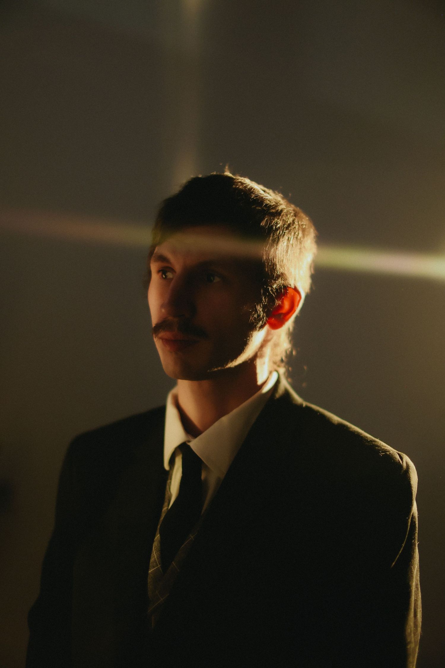 Young man with mustache in dark suit and tie illuminated by dramatic light rays in moody portrait. film, band, california