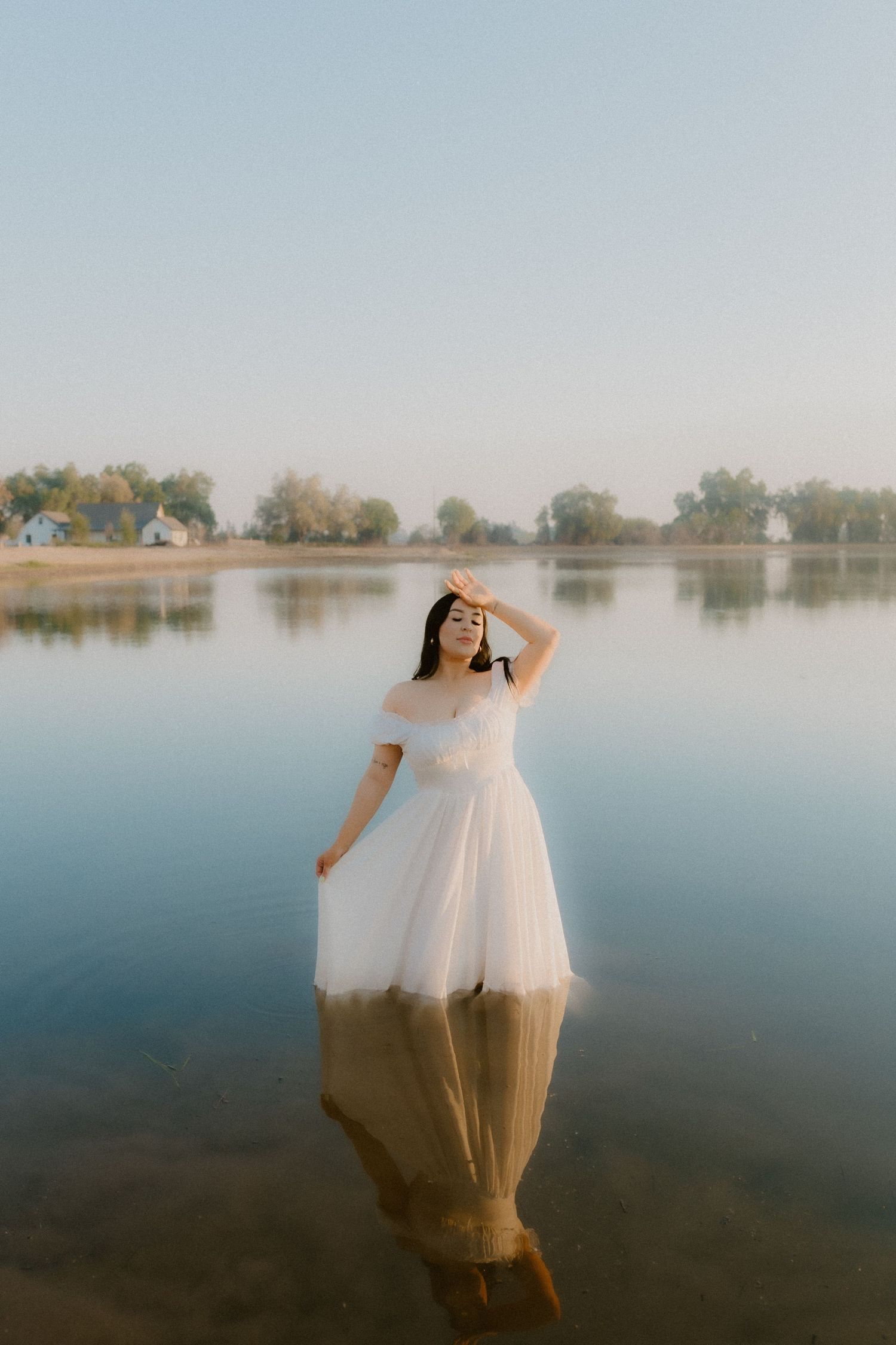 Bride in off-shoulder white tulle gown posing in shallow lake with serene reflection.