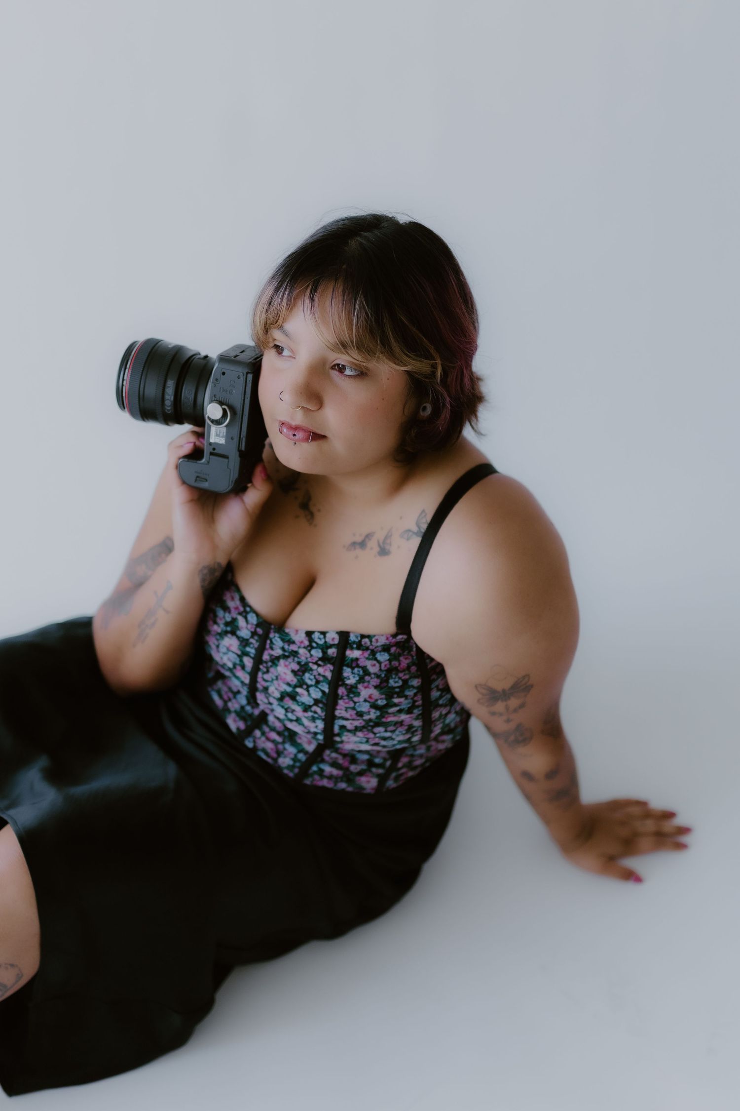 Tattooed female photographer posing with a professional camera against a white studio background.