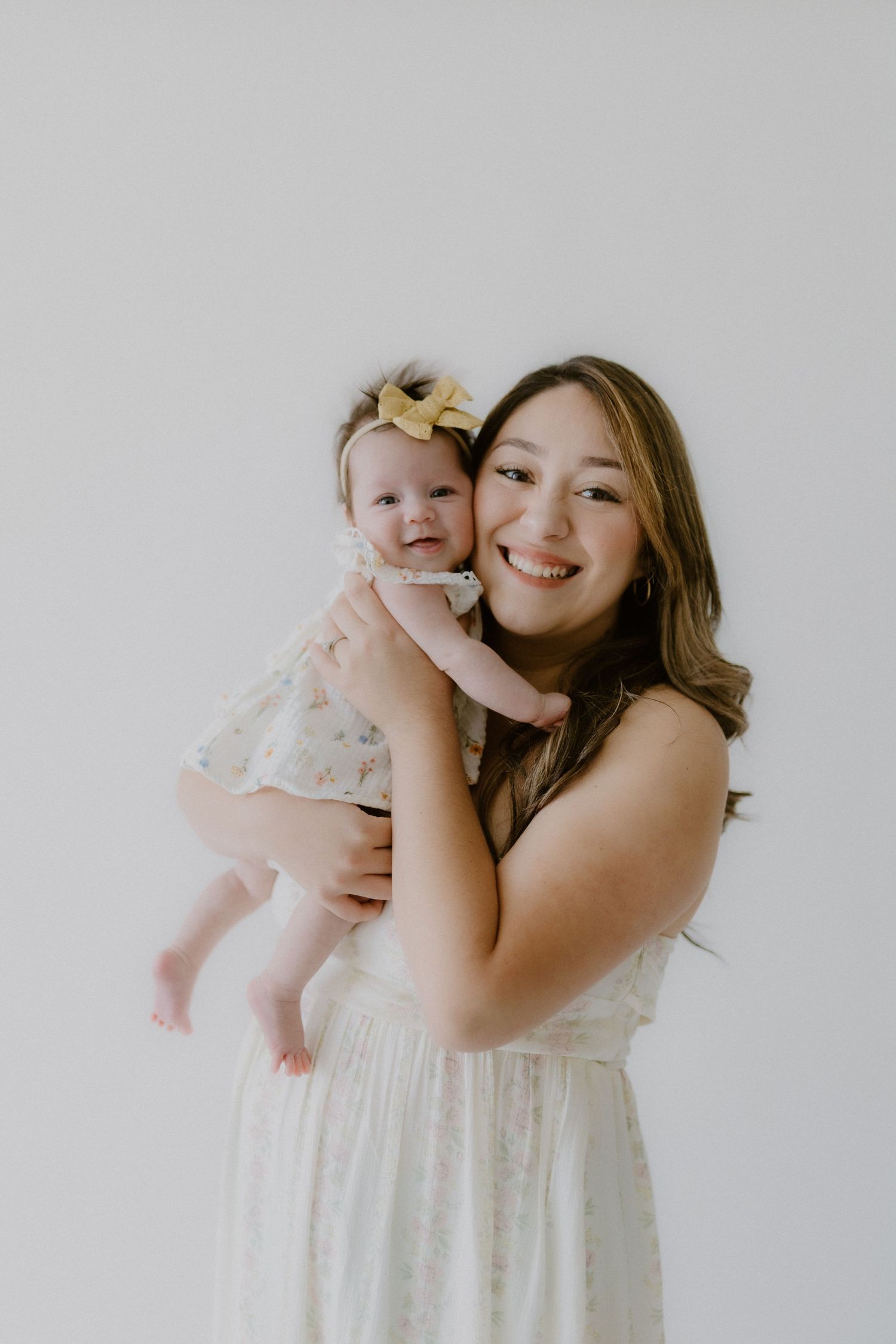 A smiling young woman in a white dress holds a happy baby girl wearing a yellow bow headband in a studio.