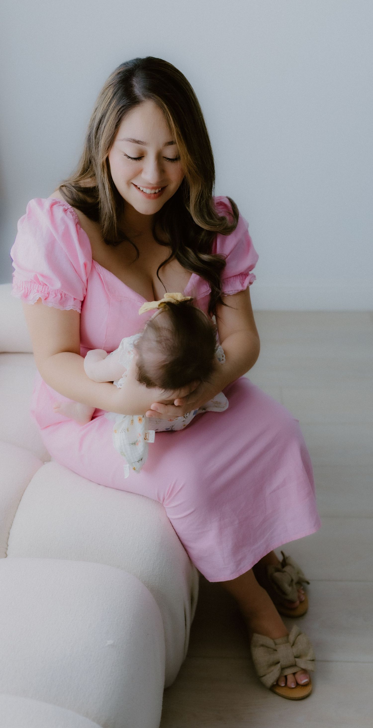 Smiling mother in a pink dress sitting on a cream sofa and cradling her newborn baby girl.