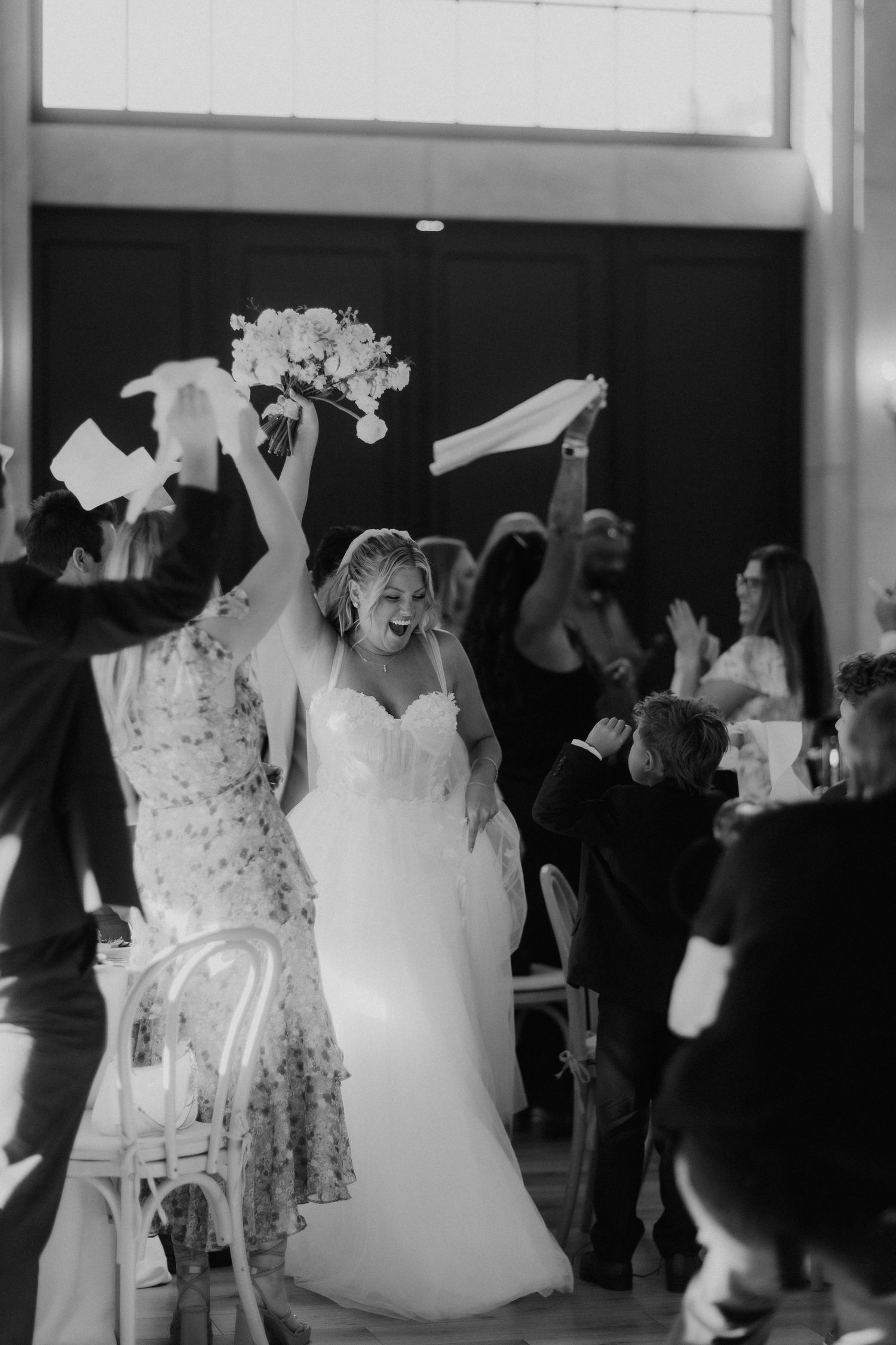 Black and white photo of radiant bride entering wedding reception with guests cheering and waving.