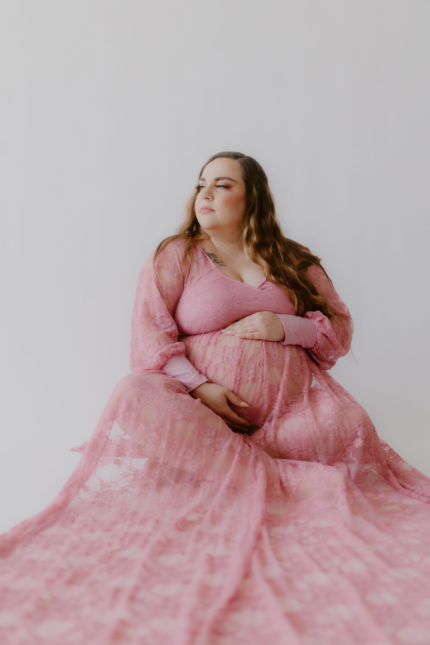 Pregnant woman in flowing pink floral gown seated gracefully against a white studio background.