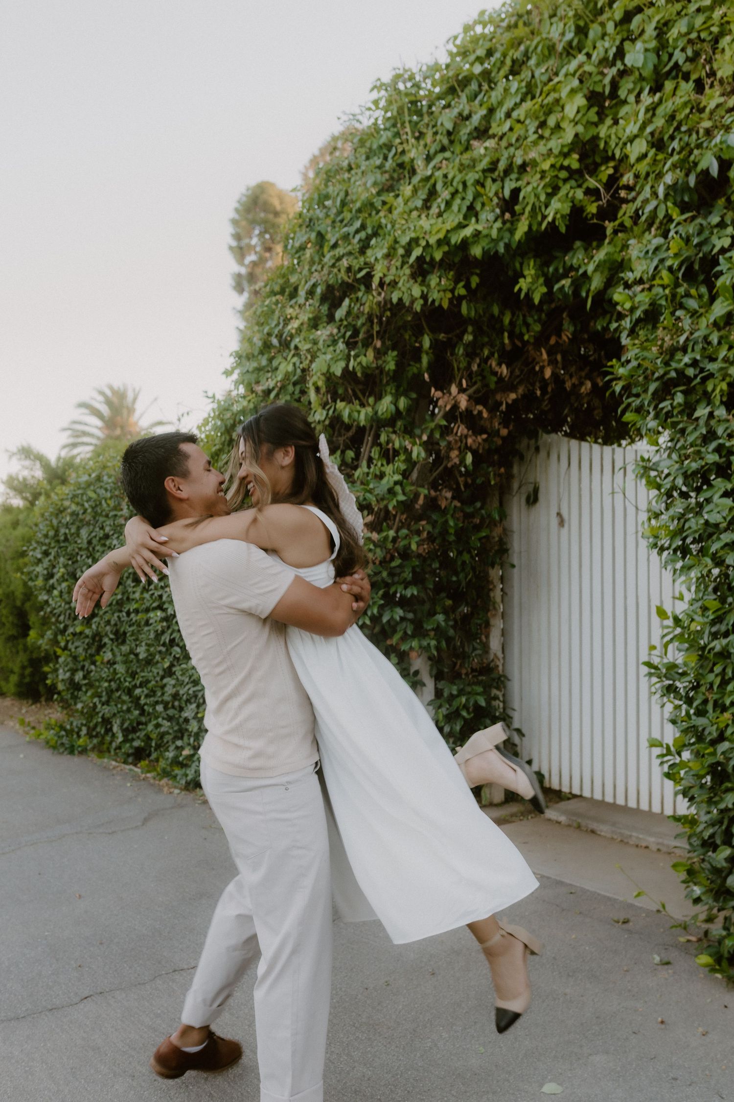 Couple in white outfits embracing and dancing near an ivy-covered white gate outdoors.