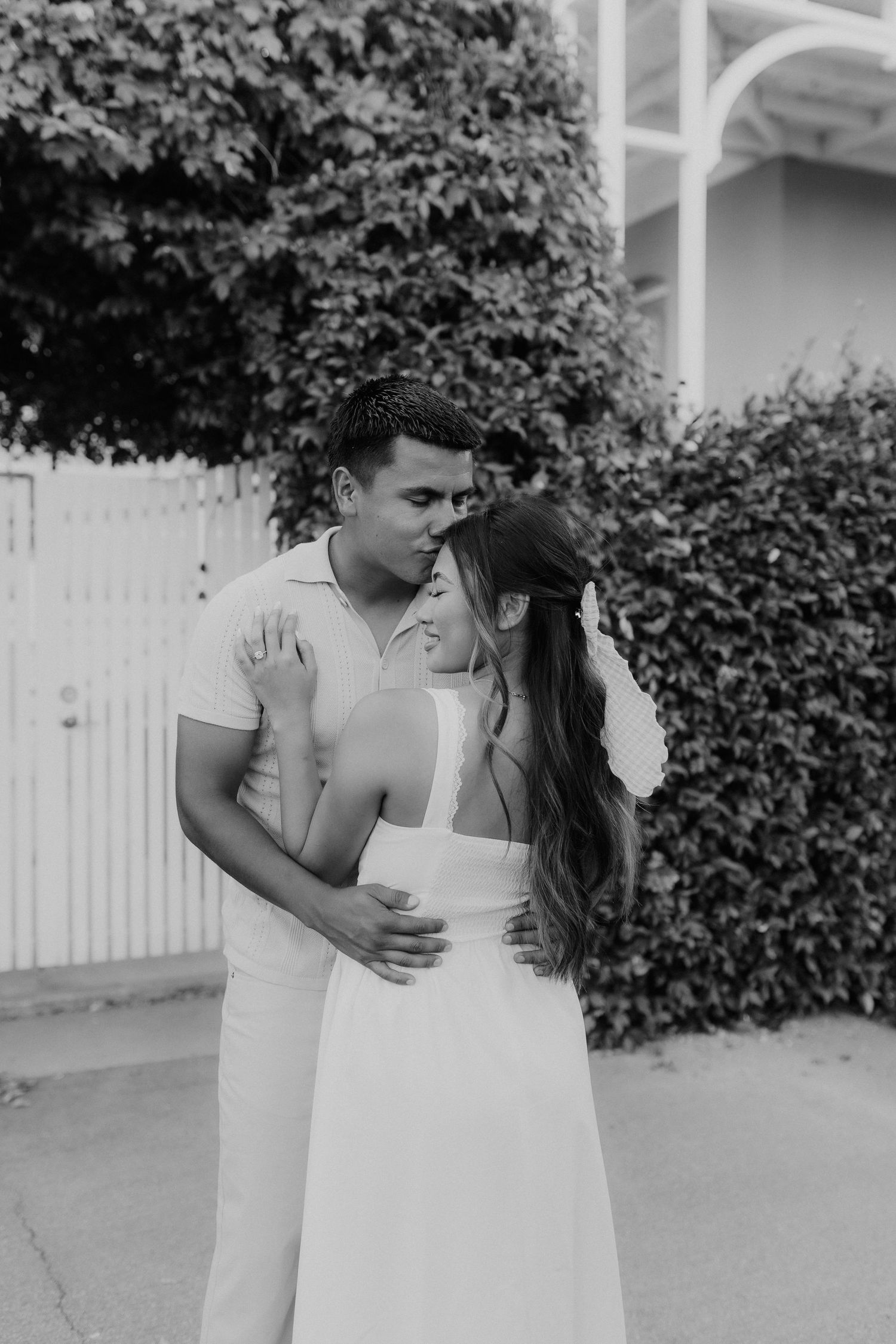 Black and white photo of an engaged couple sharing a sweet embrace near a lush ivy-covered fence.