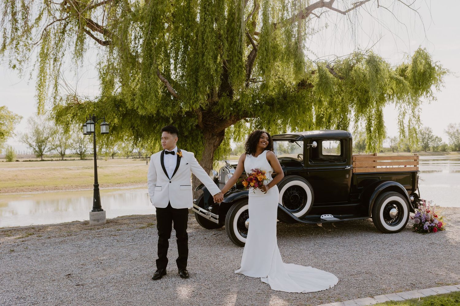 Bride and groom hold hands standing before a vintage black truck under a weeping willow tree by a lake.