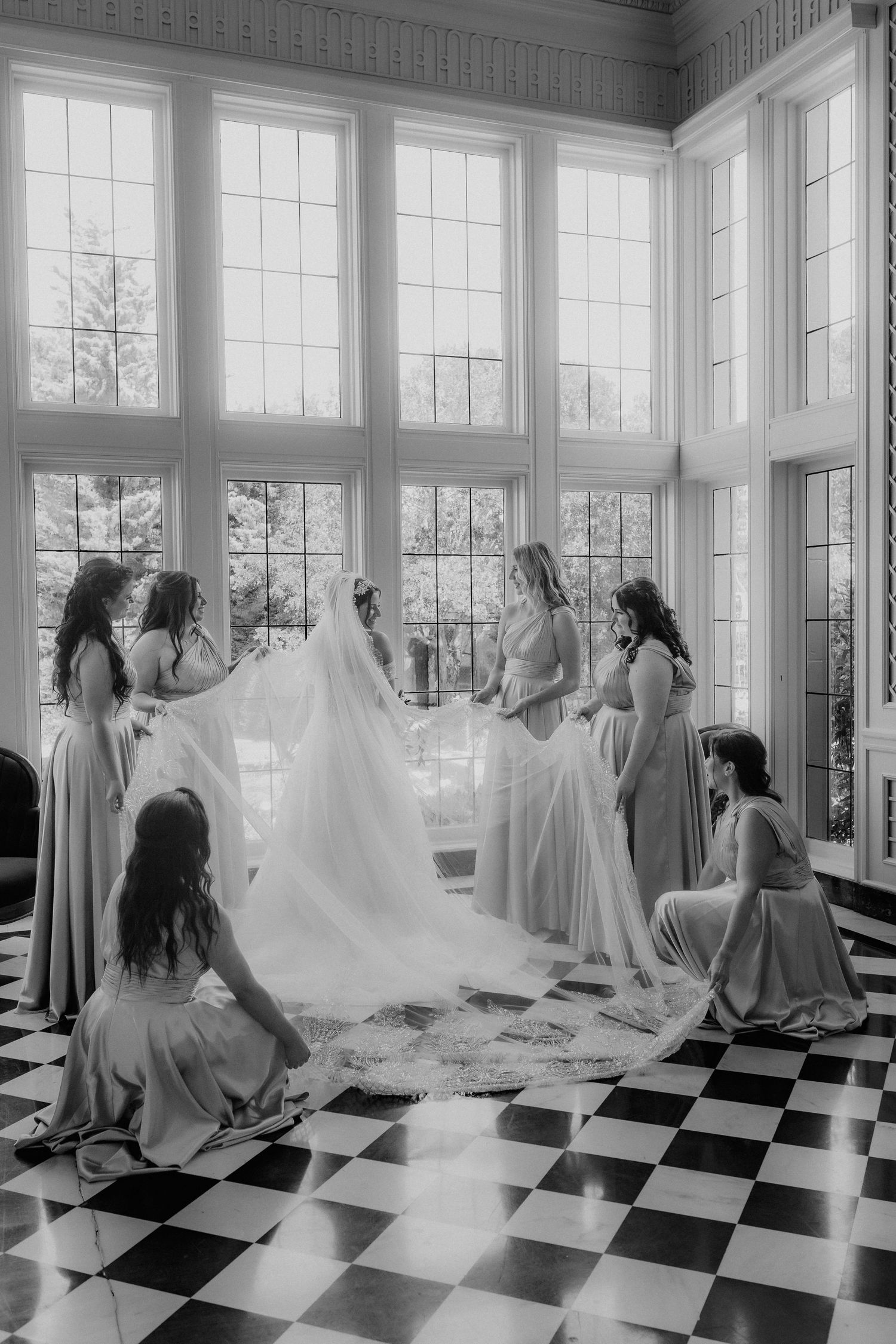 Black and white photo of bride surrounded by bridesmaids lifting her veil on checkered floor.