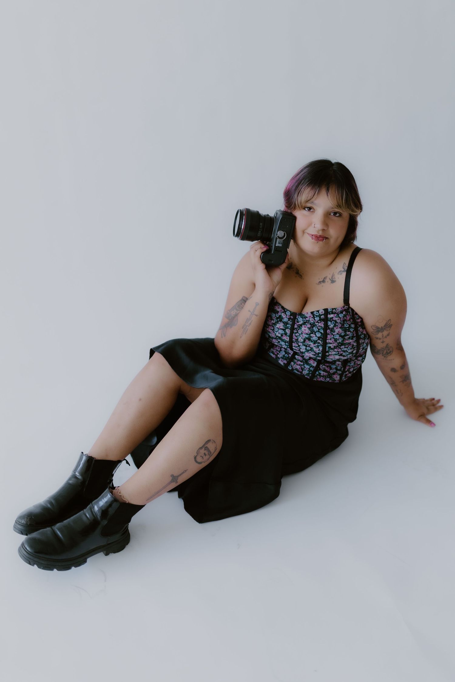 Tattooed female photographer sitting on white studio floor holding professional camera, wearing floral top and black midi skirt.