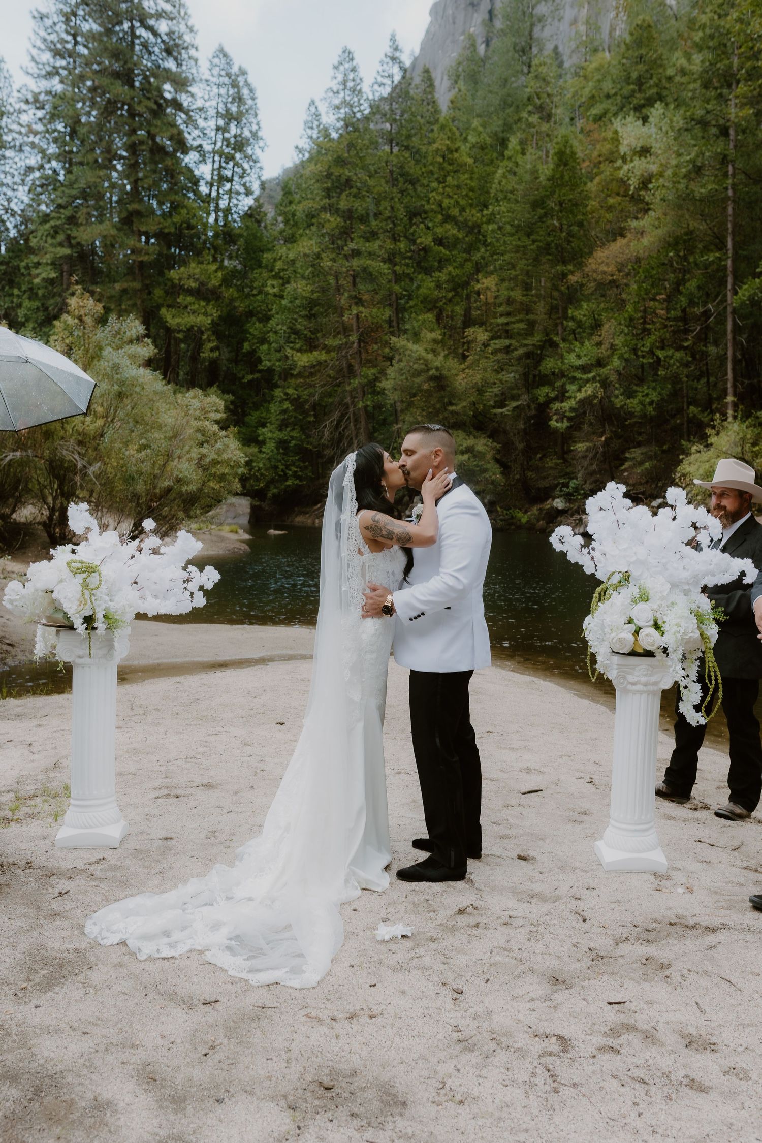 Bride and groom kissing at outdoor Yosemite wedding with white orchid floral arrangements on pedestals by a river.