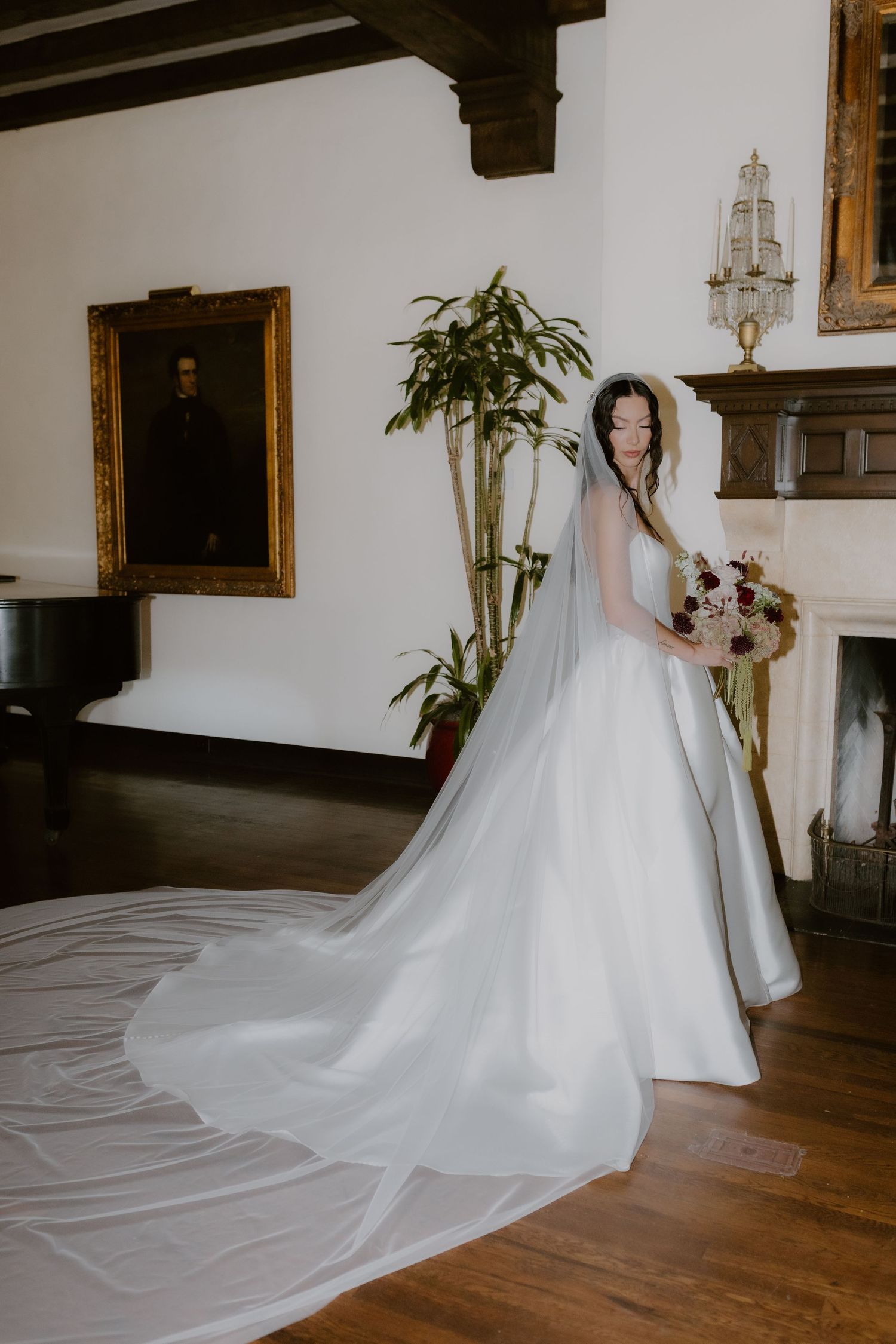Bride in strapless satin gown with cathedral veil holds bouquet beside fireplace in elegant manor.