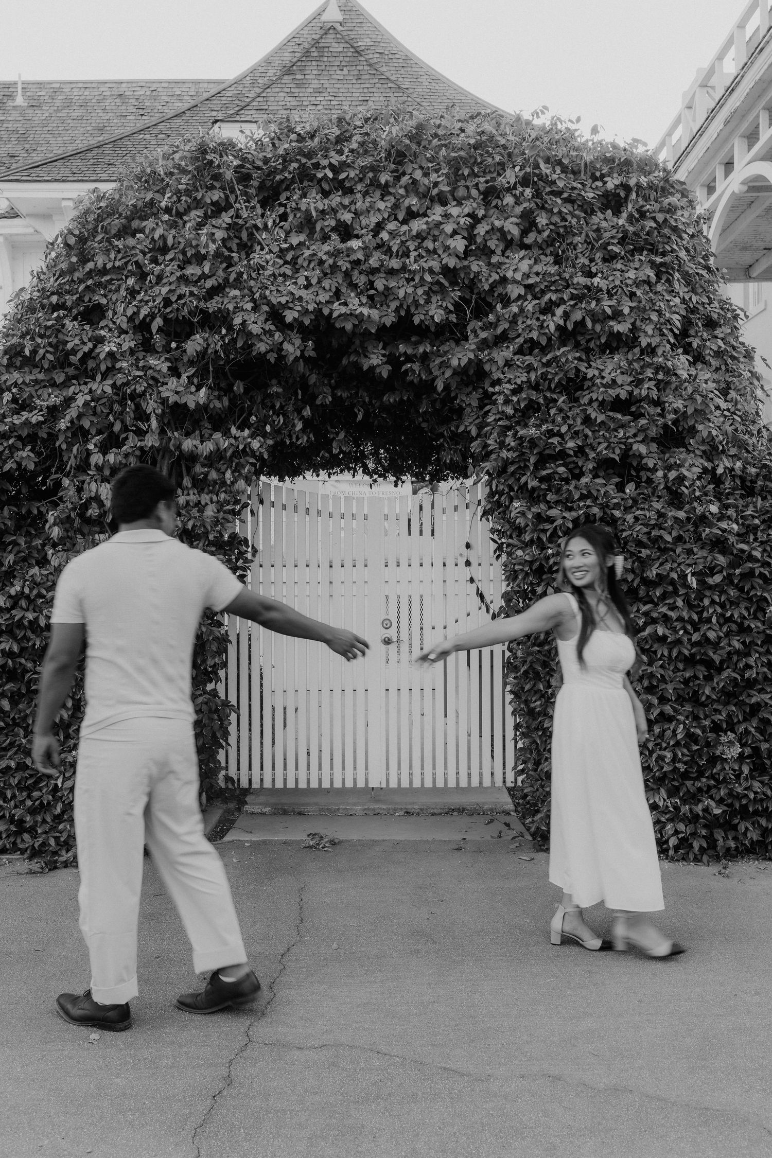 Black and white photo of couple reaching toward each other in front of ivy-covered white gate.