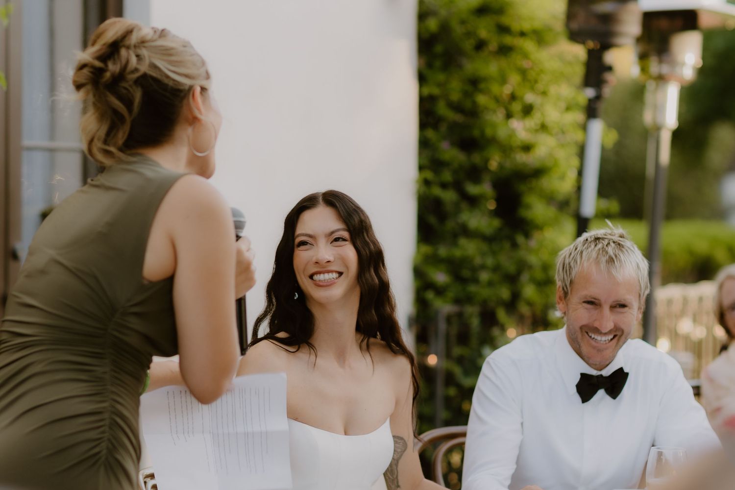 Bride laughs joyfully with groom in a white bow tie during a wedding reception speech outdoors.