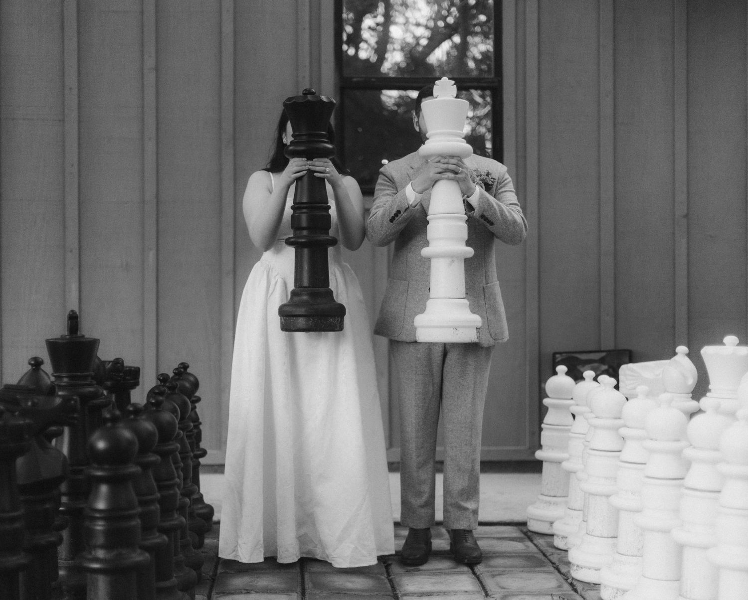 Bride and groom playfully hiding faces behind giant chess pieces at wedding reception outdoor game area. Ethereal Gardens, San Diego