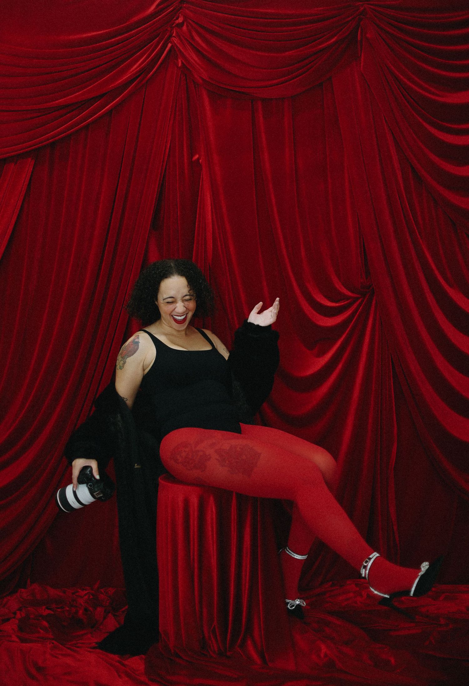 Laughing woman in red tights and black jacket poses playfully against red curtains.