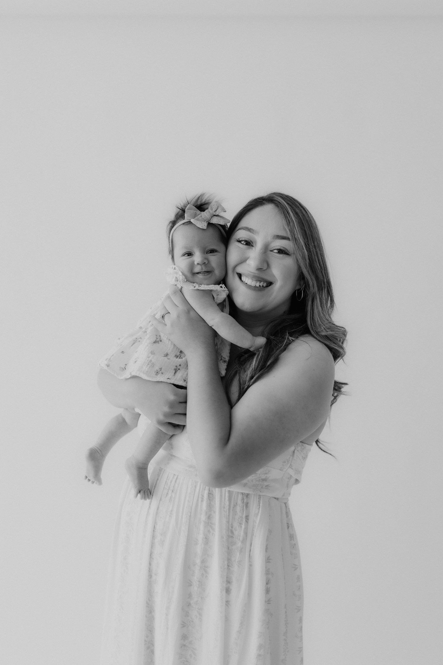 Black and white portrait of a young woman in a white dress smiling while holding a baby girl with a bow headband.