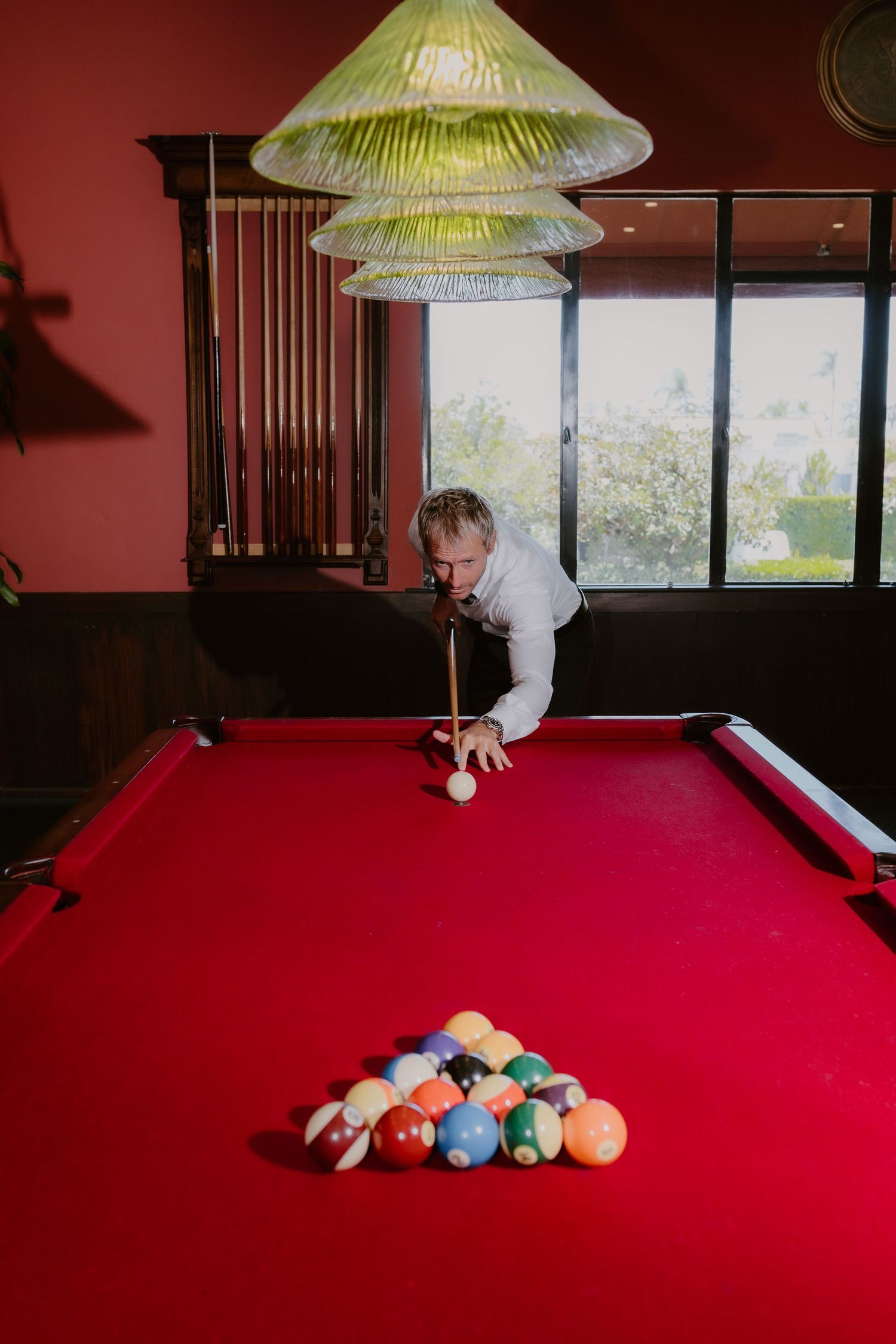 Groom leaning over red felt pool table preparing to break rack of billiard balls in elegant game room.